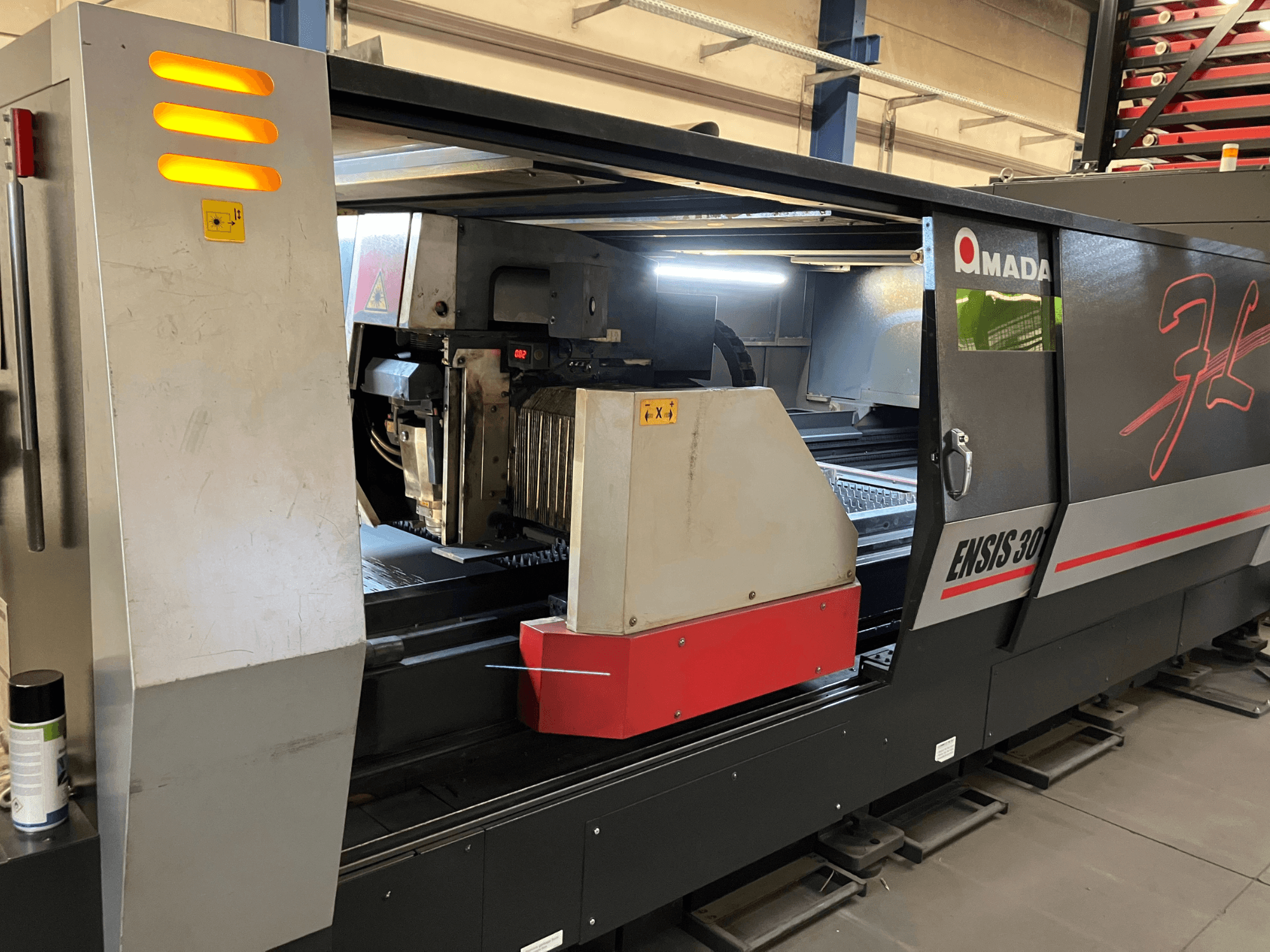 AMADA ENSIS 3015 AJ Faserlaserschneidmaschine, Vorderansicht, zeigt Schneidkopf und Bedienelemente in einem Metallrahmen.