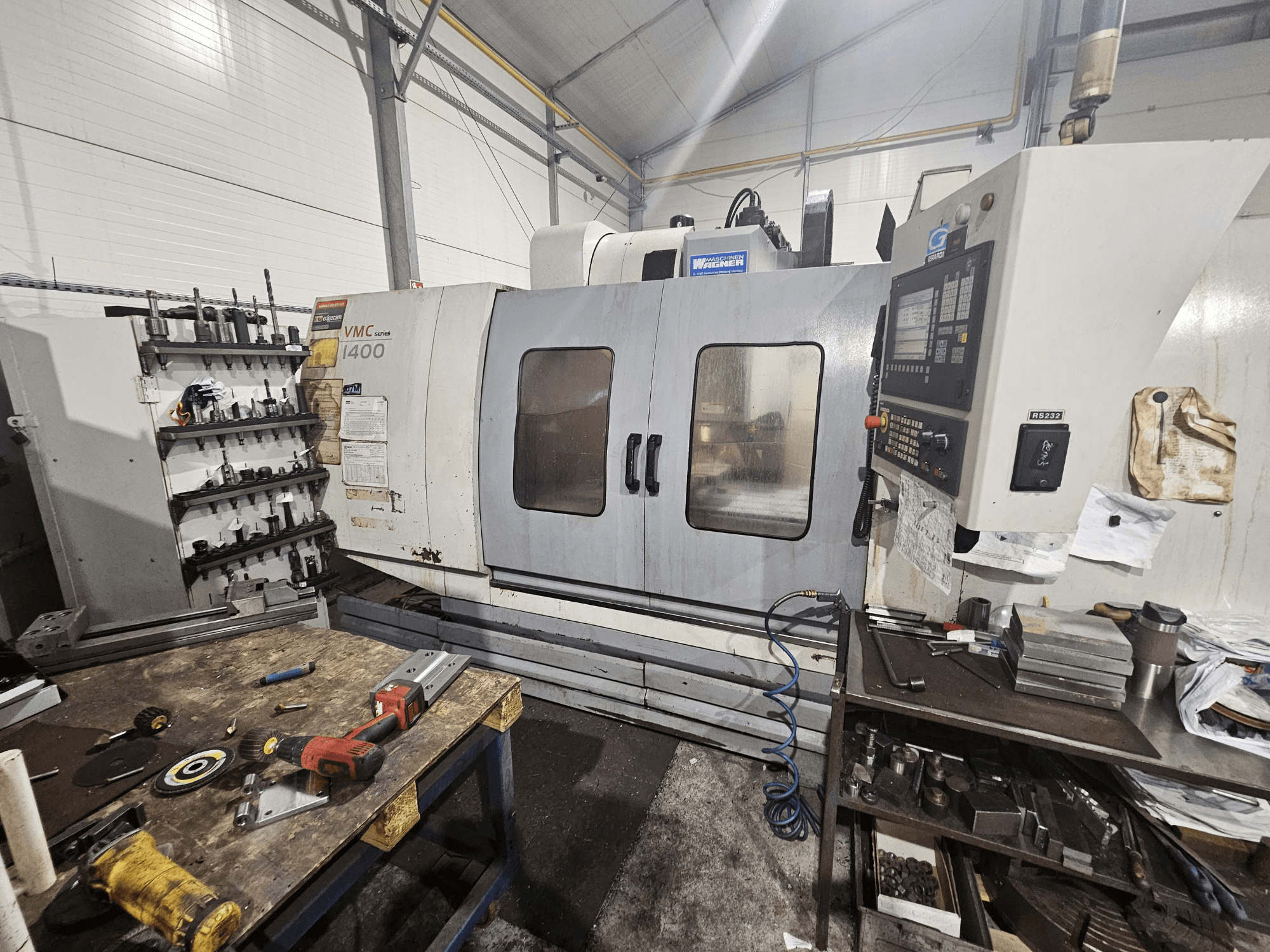 VMC 1400 CNC-Fräsmaschine in einer Werkstatt, Vorderansicht; Werkzeuge und Materialien liegen verstreut auf der Werkbank daneben.