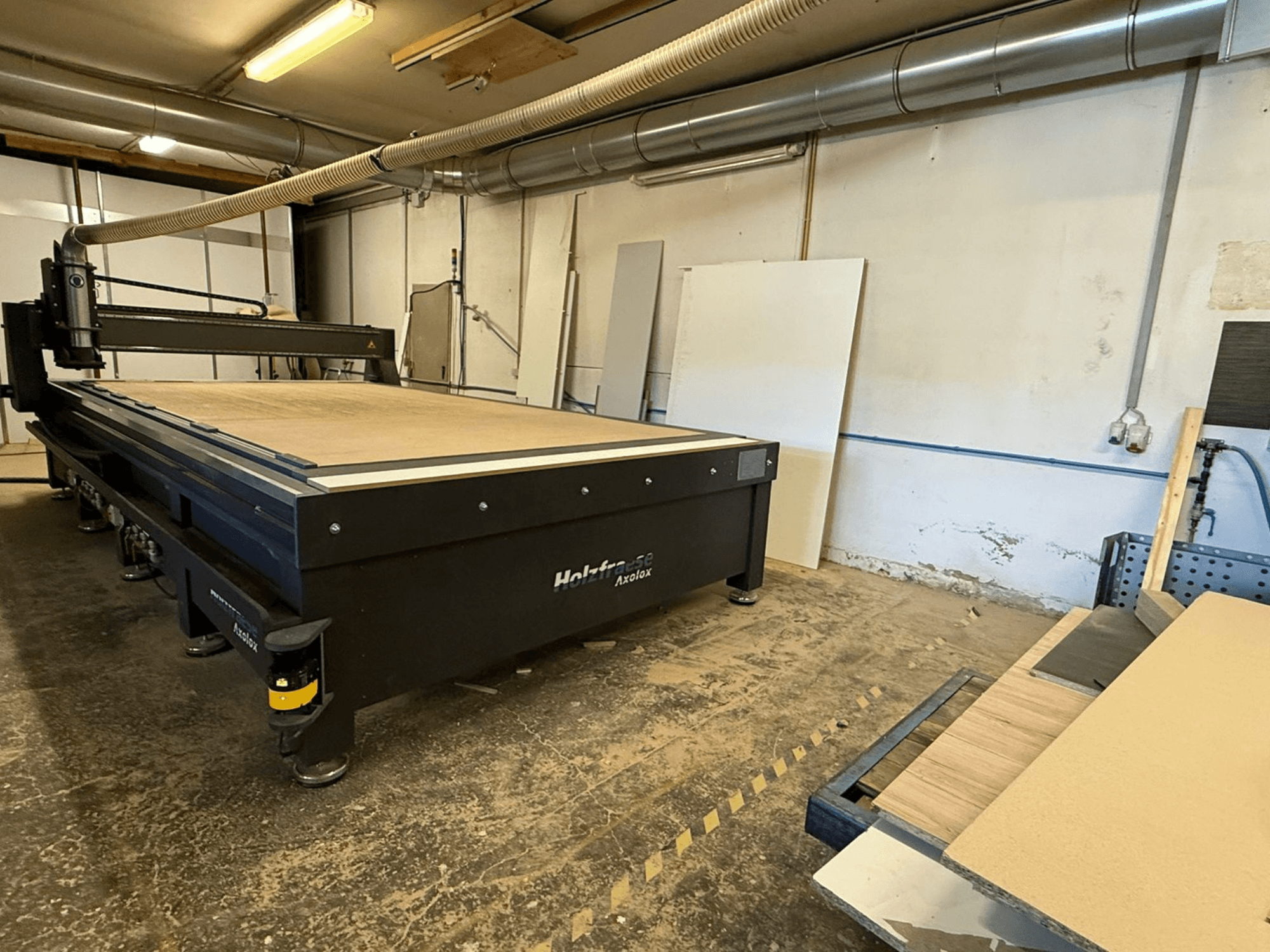 Holz-Her Axos 5 CNC-Oberfräse in der Werkstatt, Vorderansicht mit Arbeitstisch, Absauganlage und Holzplatten daneben.