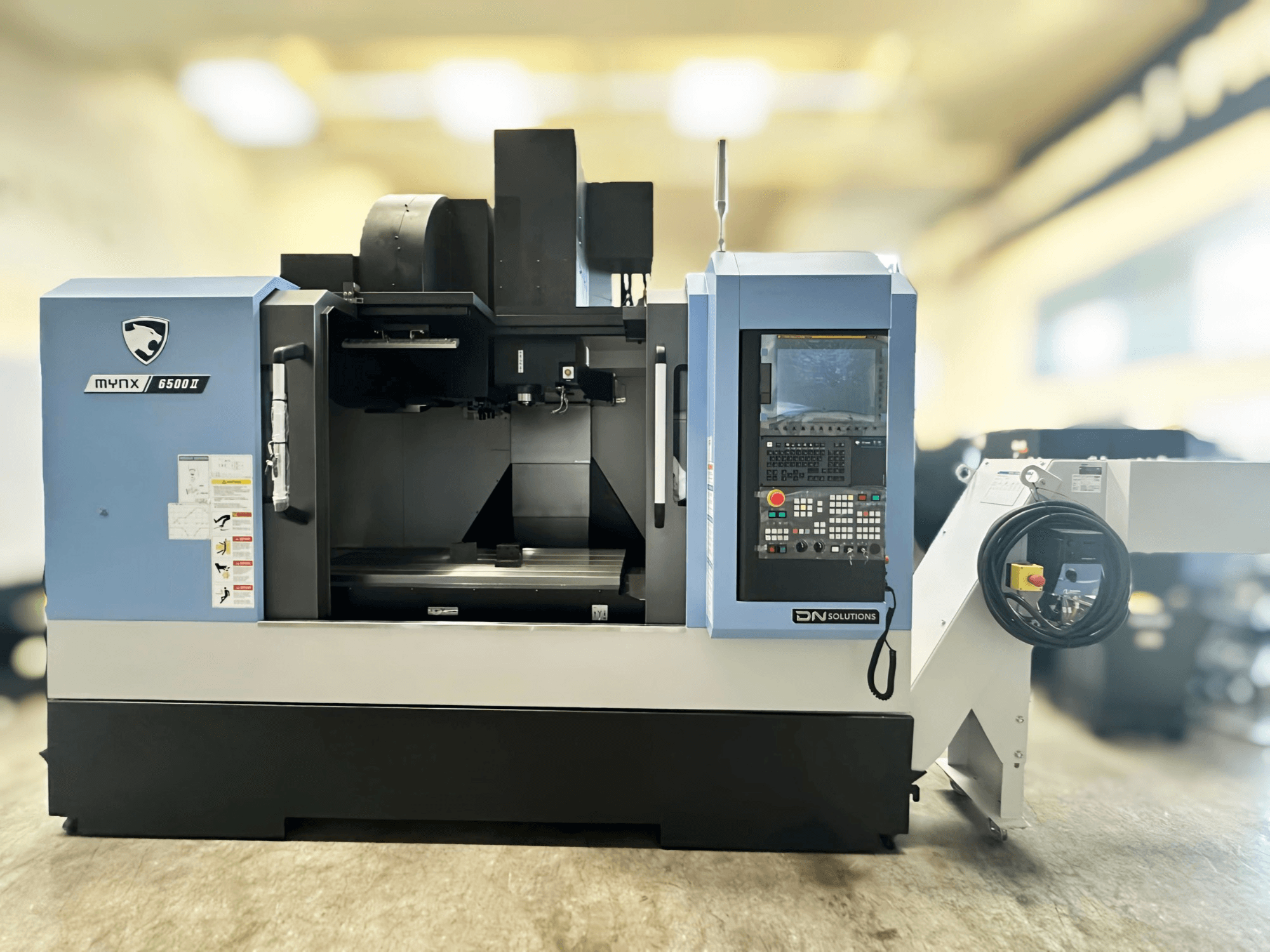 Vertikales Bearbeitungszentrum „DN SOLUTIONS MYNX 6500 II“ in der Vorderansicht, mit Bedienfeld und Bedienoberfläche.