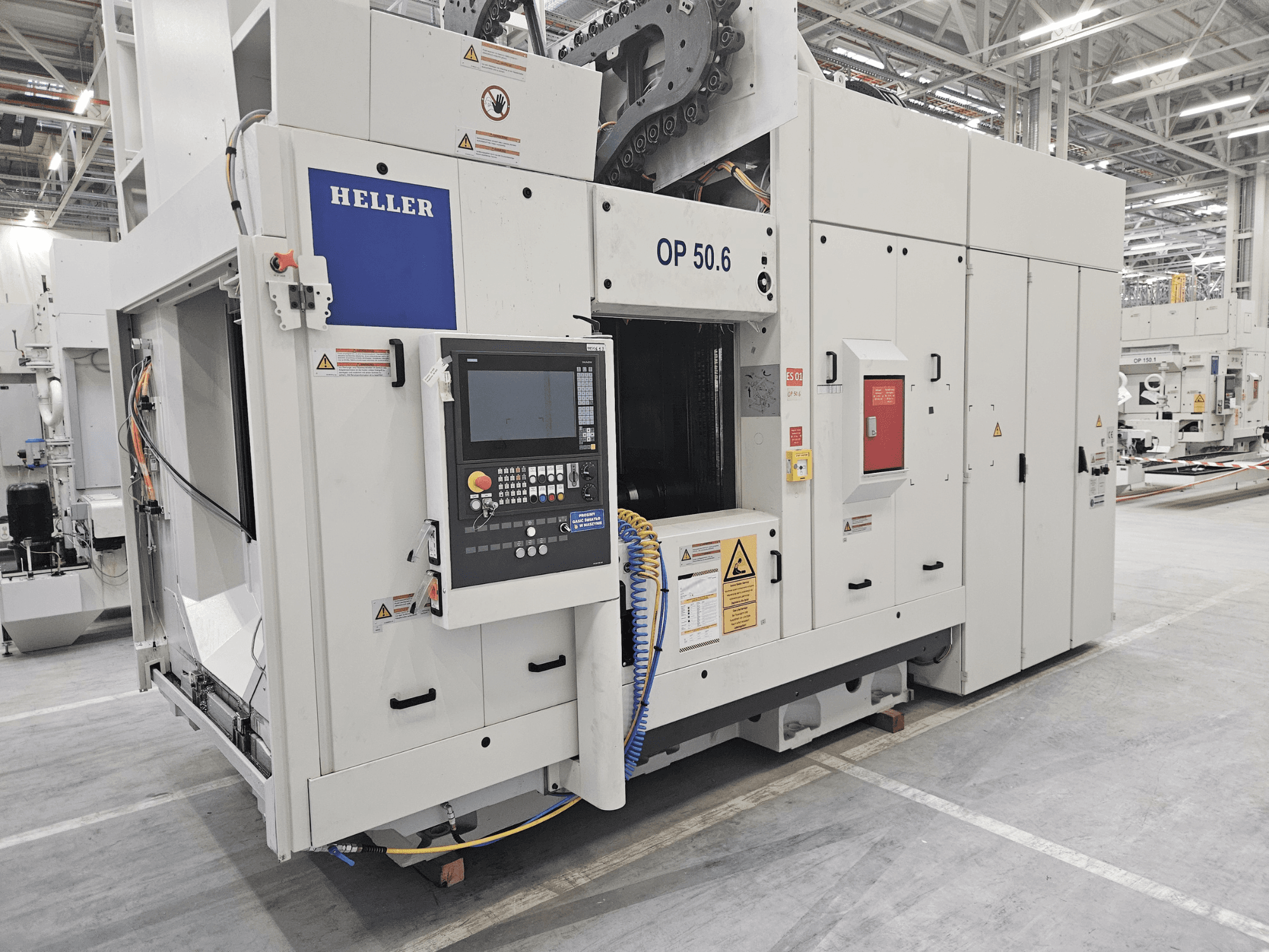 Heller OP 50.6 Bearbeitungszentrum, Vorderansicht, mit Bedienfeld, Warnschildern und Schläuchen in einer Fabrikumgebung.