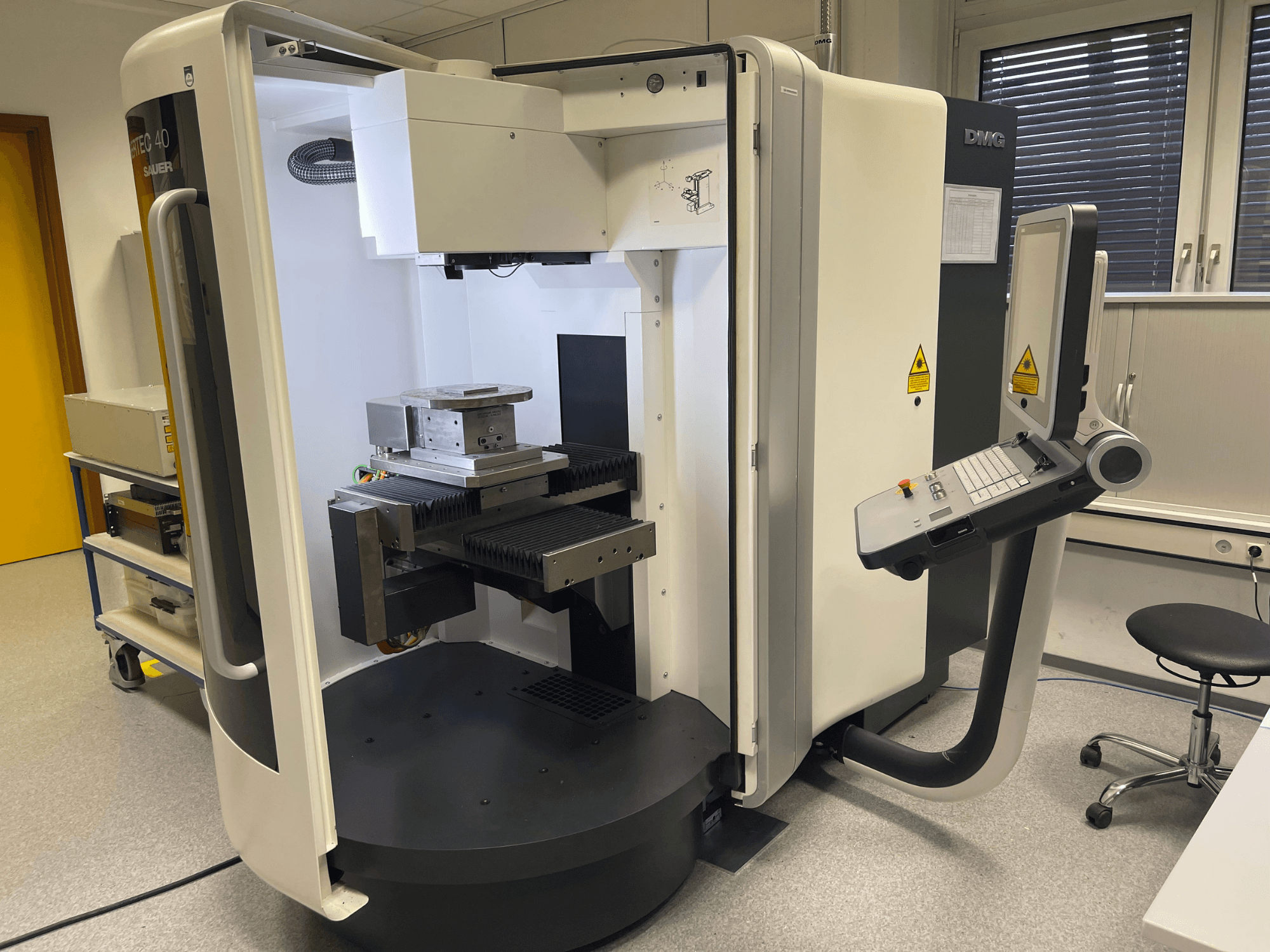 DMC 40 Maschine, Seitenansicht, mit Bedienfeld und Arbeitstisch mit einem Metallteil, in einer Laborumgebung.