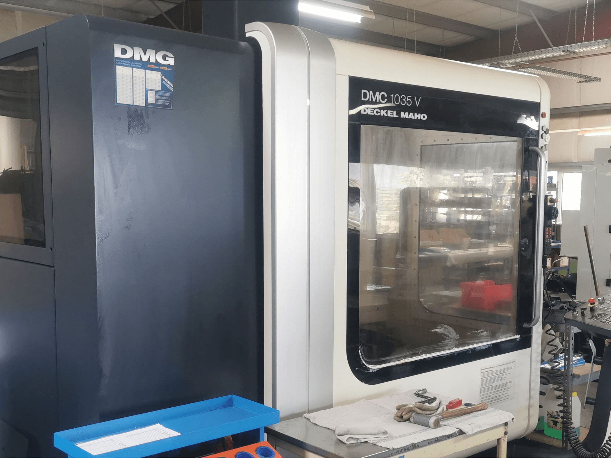 DMG DMC 1035 V Fräsmaschine in einer Werkstatt, Vorderansicht mit blauem Werkzeugkasten und Handschuhen auf der Oberfläche.