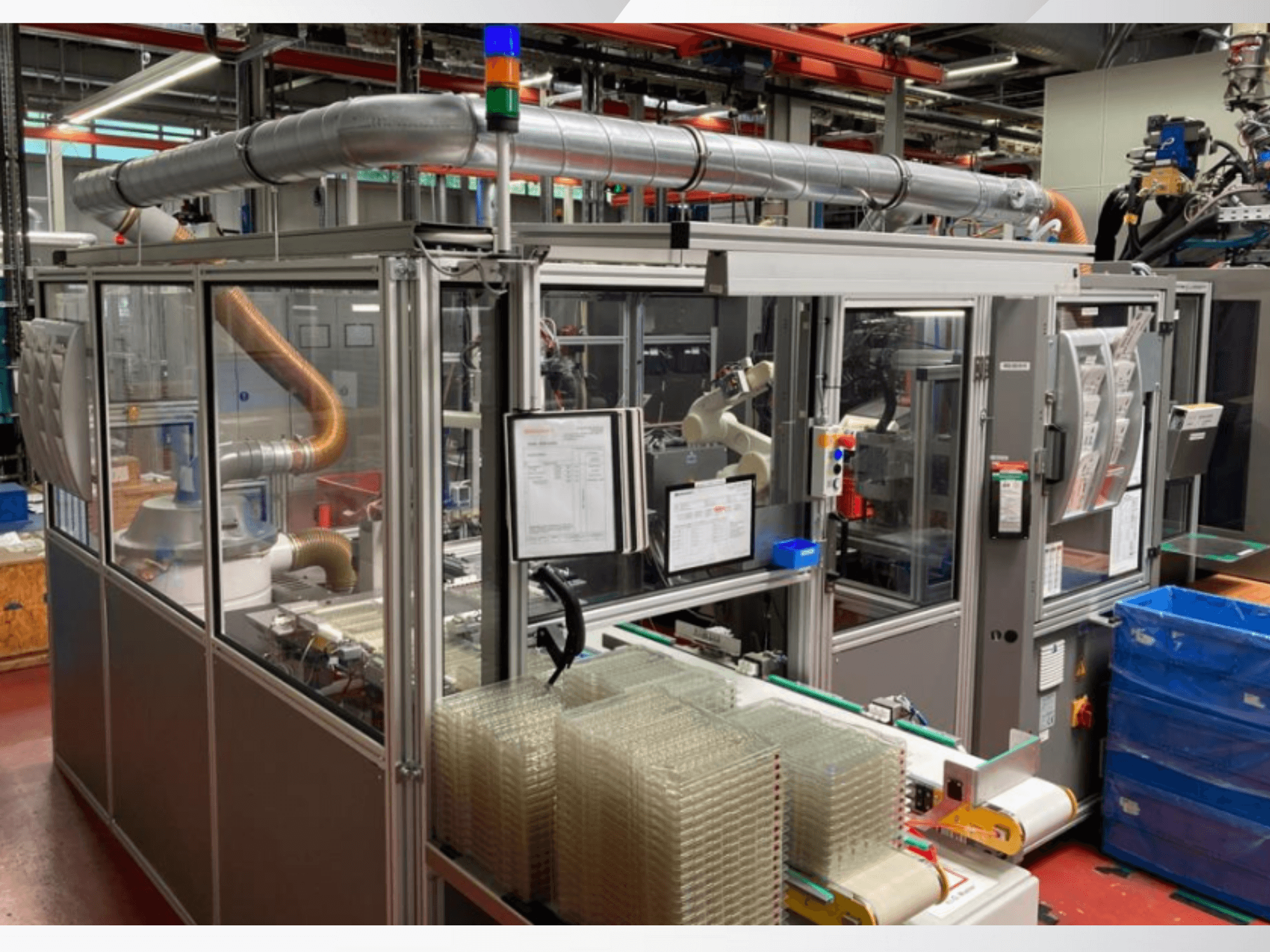 Automatisierte Montagemaschine mit Roboterarm, transparenten Seitenwänden und gestapelten Behältern mit durchsichtigen Artikeln, Vorderansicht.