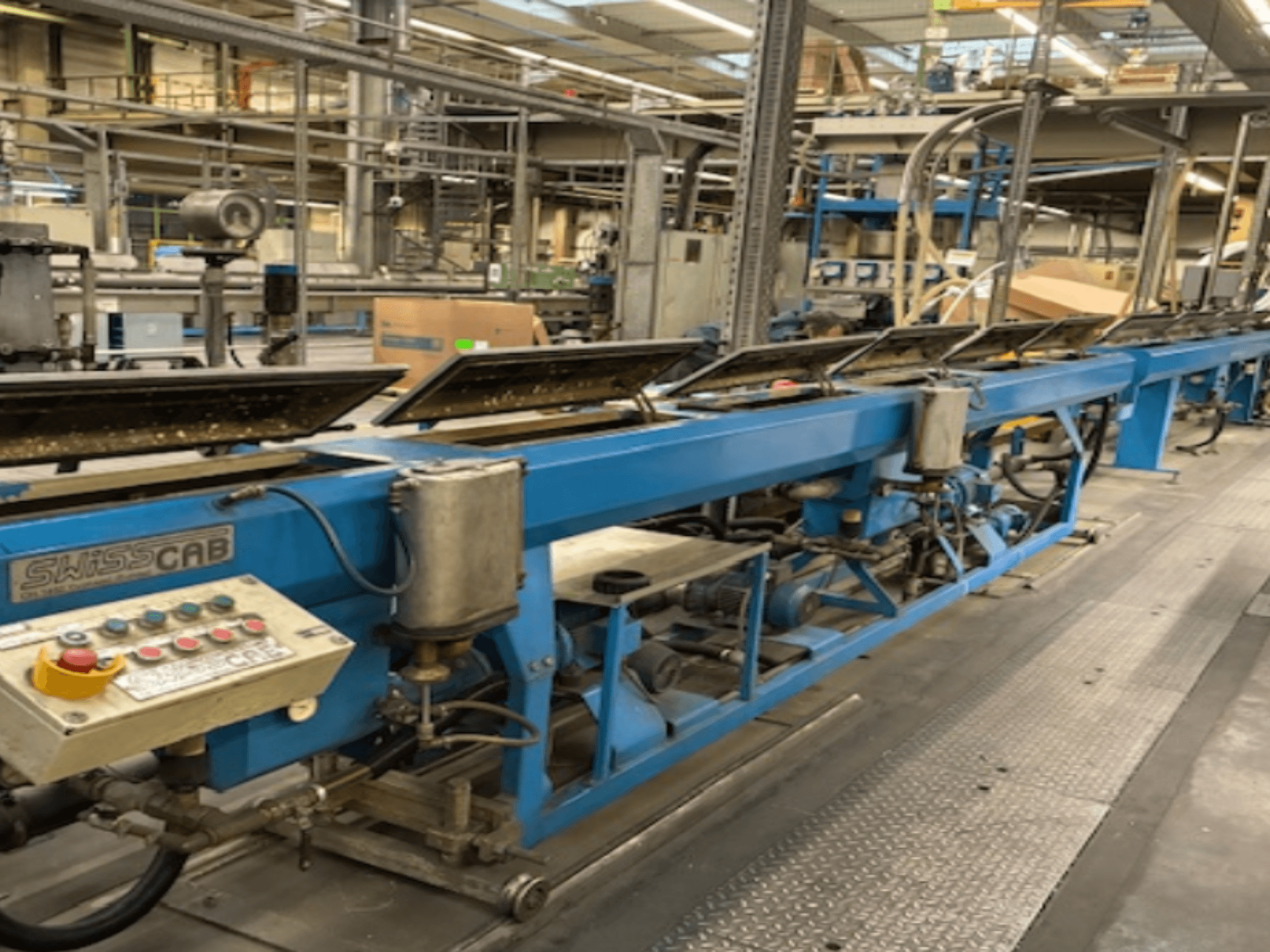 Blaue SwissCab-Industriemaschine in einer Fabrikumgebung, mit Bedientasten und einem Fördersystem, Seitenansicht.