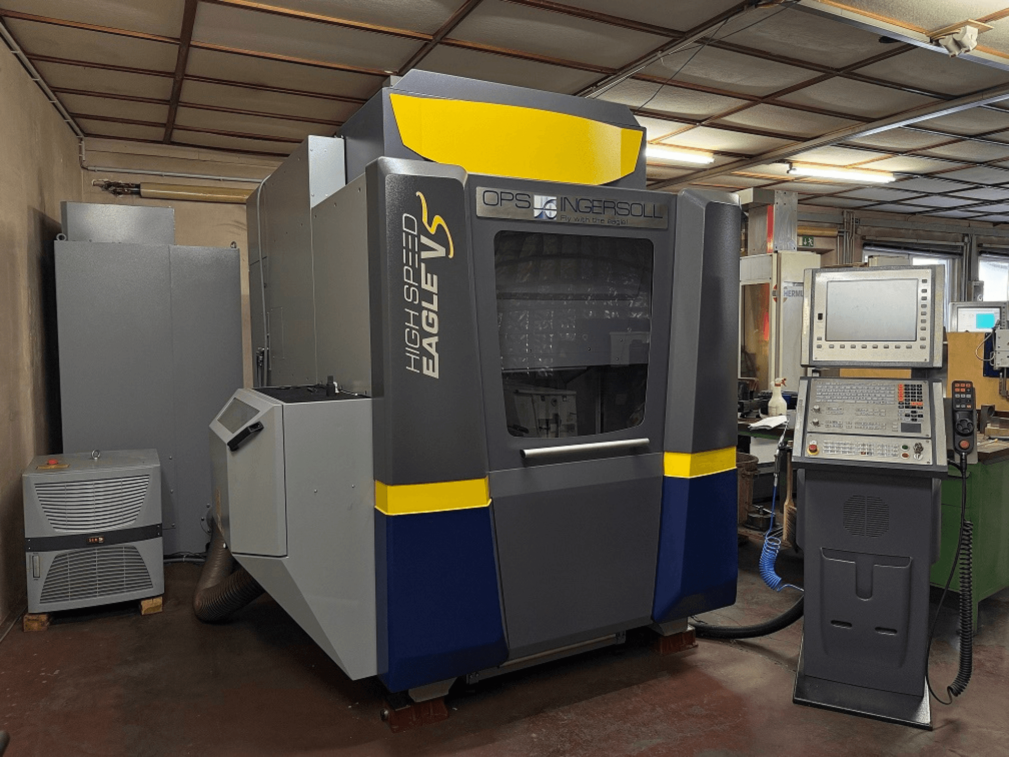 Hochgeschwindigkeitsmaschine Eagle V von OPS Ingersoll, Seitenansicht, mit Bedienfeld und angrenzenden Geräten in einer industriellen Umgebung.
