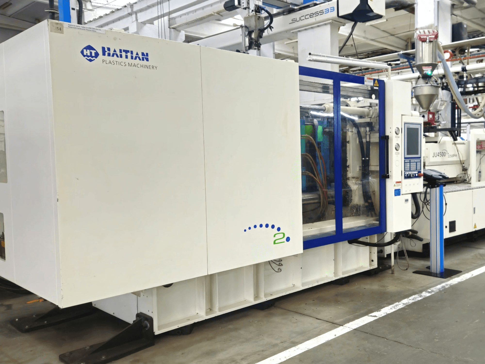 Modell von Haitian Plastics Machinery, Vorderansicht, zeigt eine Maschine mit Bedienfeld und sichtbaren Schläuchen in einer industriellen Umgebung.