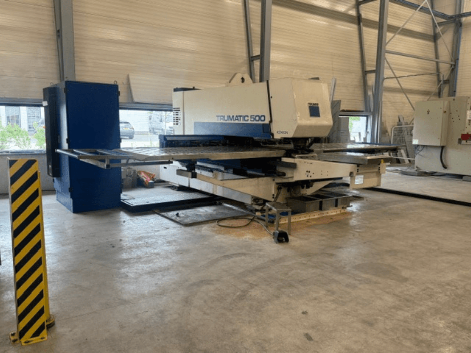 CNC-Stanzmaschine TRUMPF Trumatic 500 in einer Werkstatt, Seitenansicht mit Maschinenanlage und Sicherheitspfosten mit gelben Streifen.