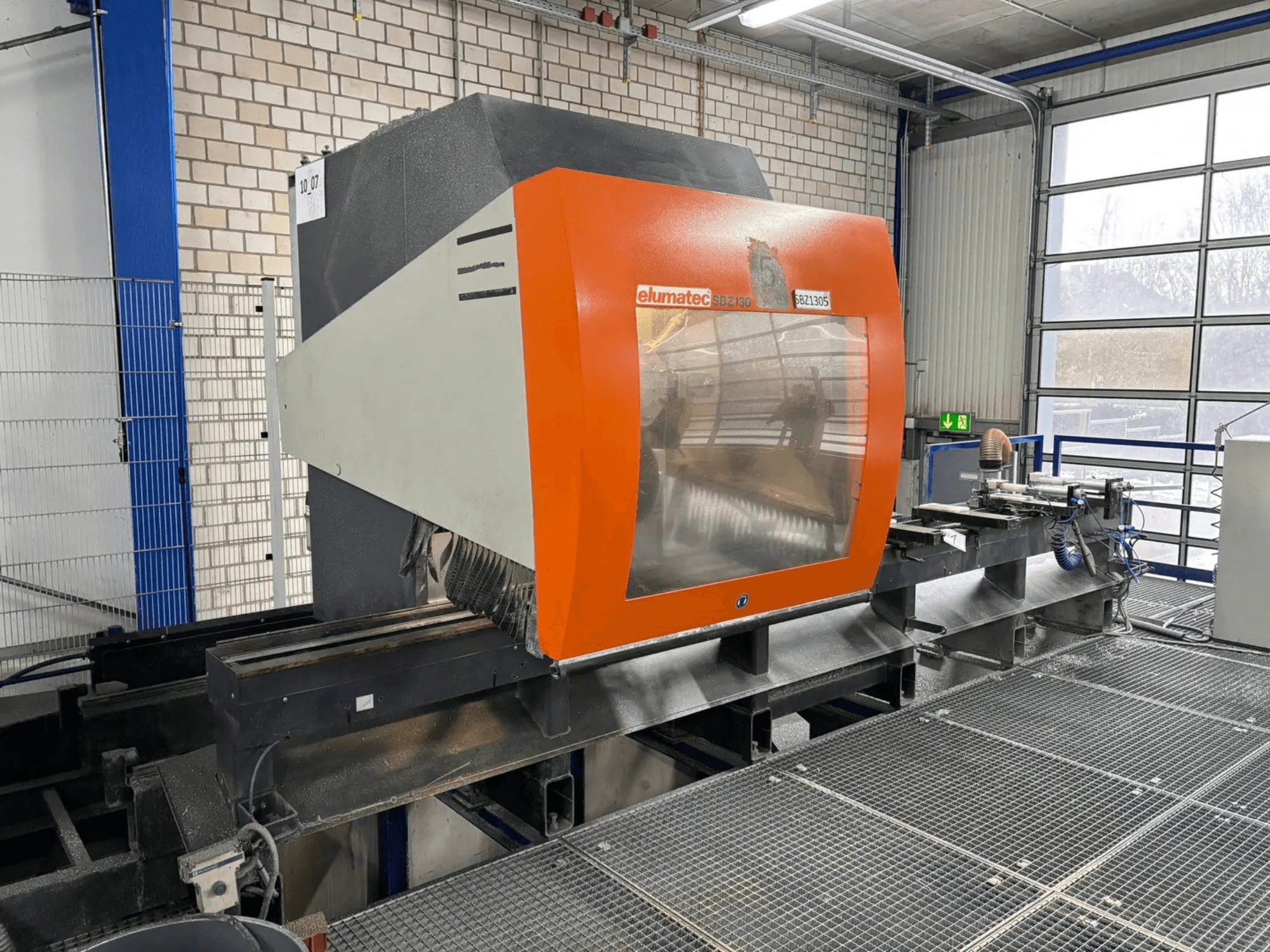 Elumatec SBZ 130 Vertikal-Bearbeitungszentrum, Frontansicht, mit orange-grauem Gehäuse und transparenter Scheibe.