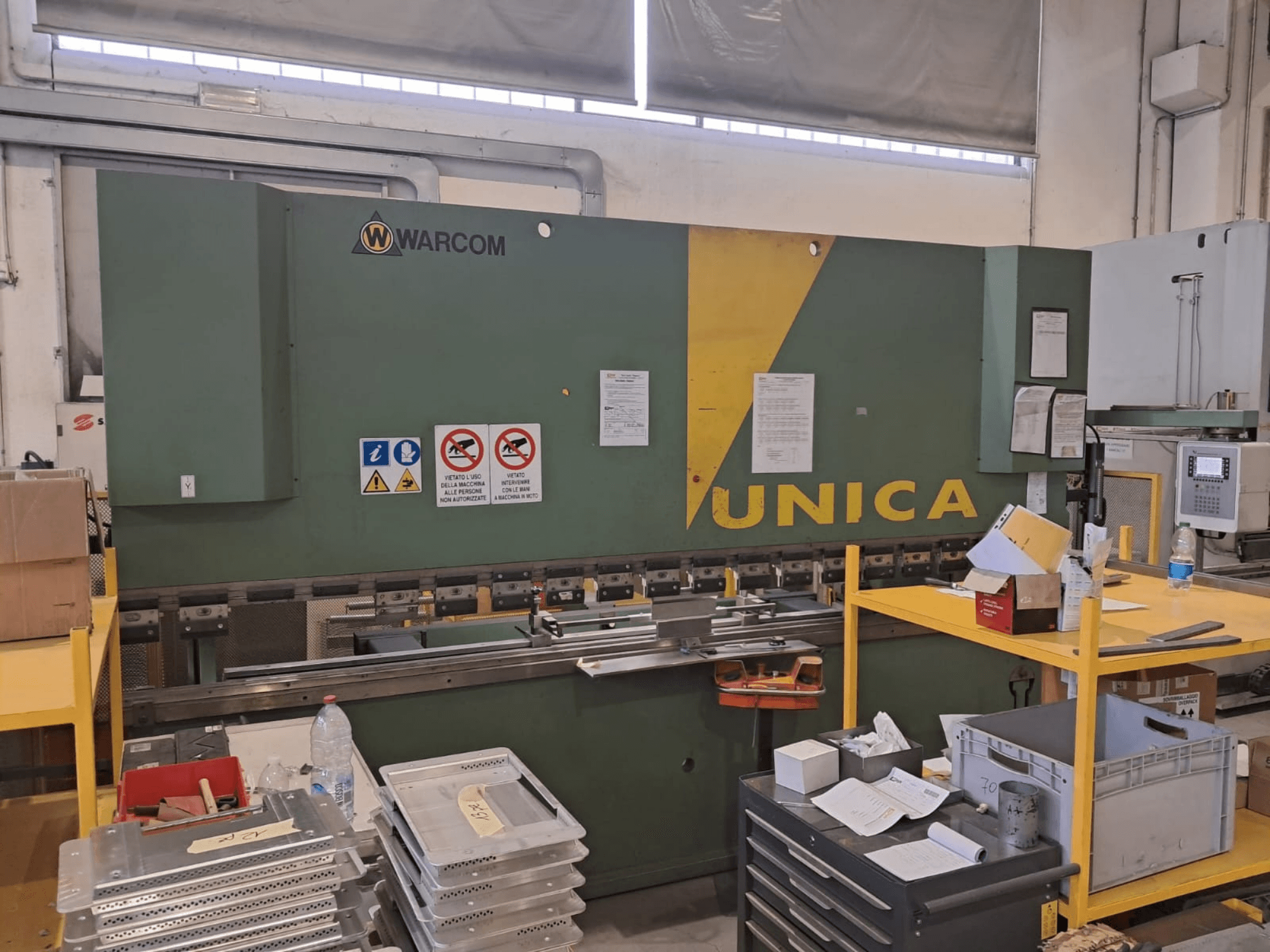 Hydraulikpresse Warcom UNICA 100-40, Vorderansicht, grünes Gehäuse mit gelben Akzenten, Sicherheitsschilder sichtbar, Werkzeuge und Materialien auf dem Tisch.