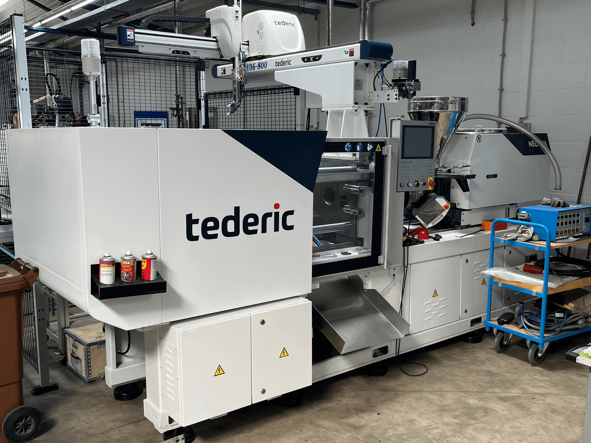 Hydraulische Spritzgießmaschine TEDERIC NEO.E55/e110h in einer Werkstatt, Seitenansicht mit Bedienfeld und angrenzenden Werkzeugen.