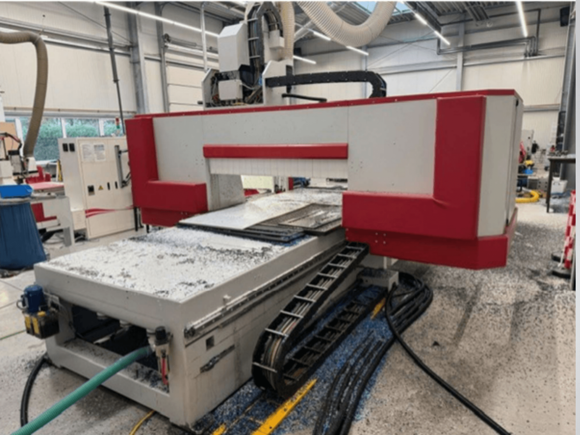 Rote und graue CNC-Maschine in einer Werkstatt, Seitenansicht mit Materialtisch, Kühlmittelschlauch und Sägemehl auf dem Boden.