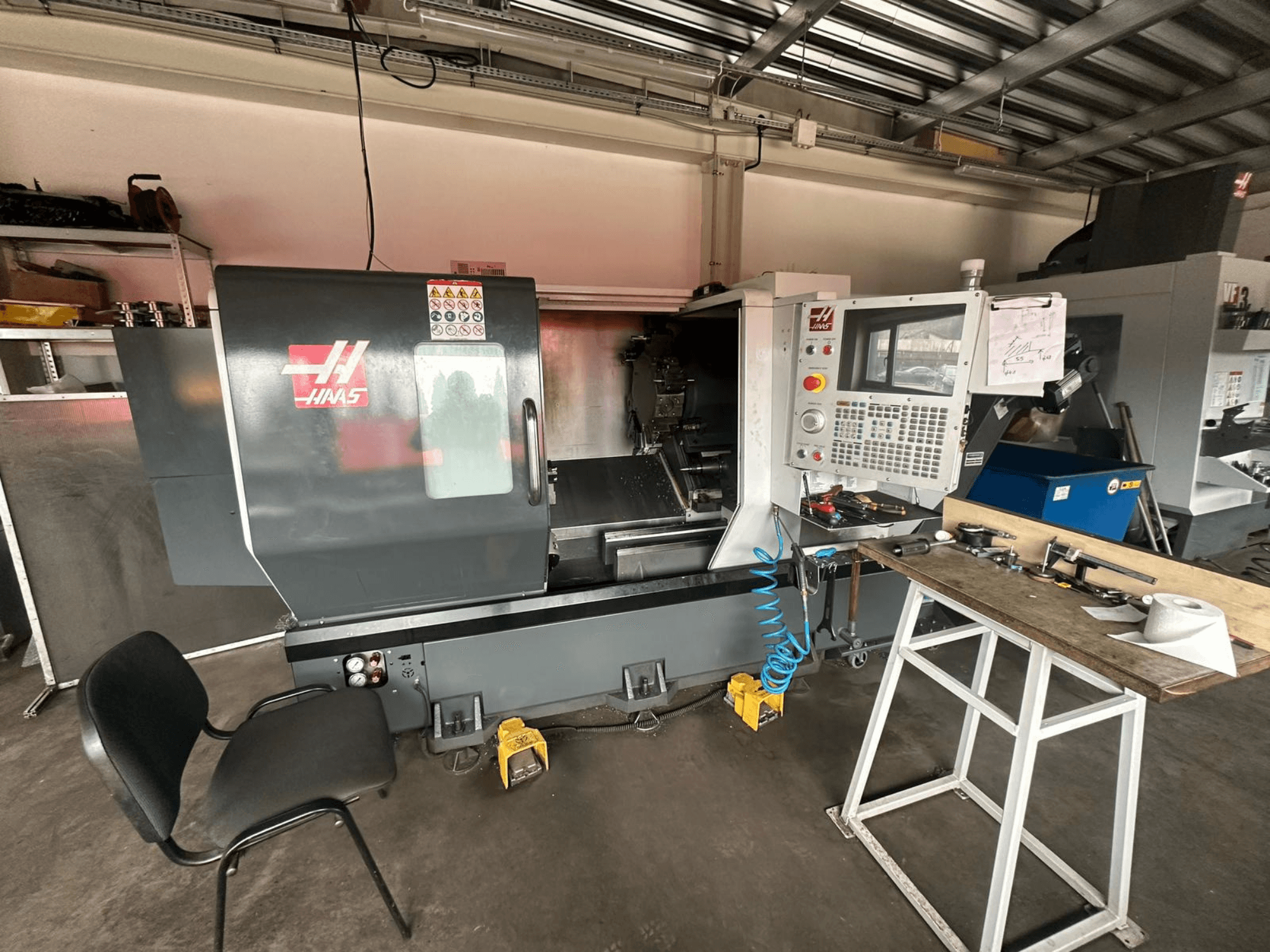 Haas CNC-Drehmaschine (Modell ST-10) in einer Werkstatt, Seitenansicht mit Bedienelementen, Werkzeugen auf einer Werkbank und einem Stuhl in der Nähe.