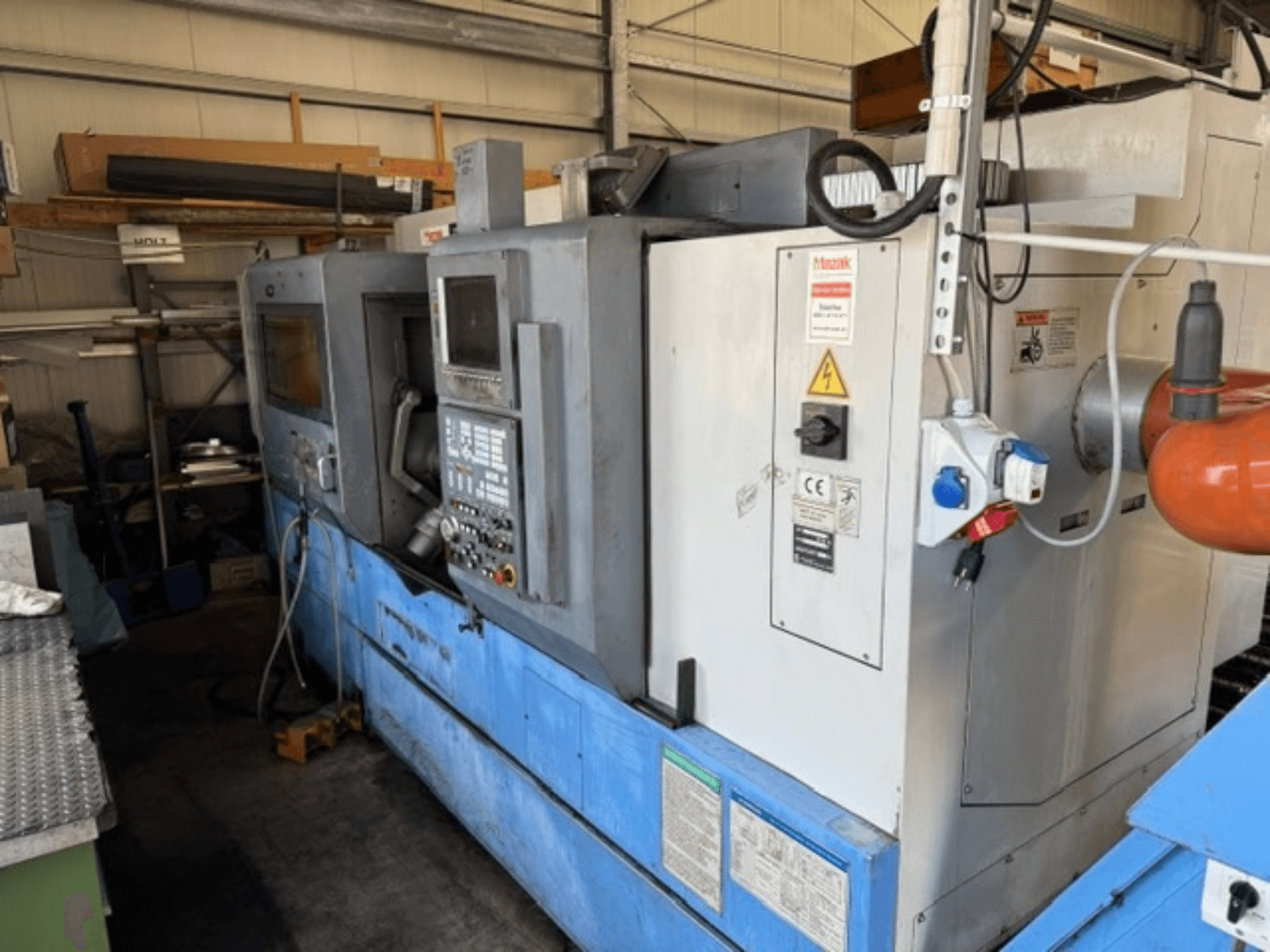 Mazak SQT-30M * 1000 Dreh-Fräszentrum, Seitenansicht mit Bedienfeld, blauem Sockel und umgebenden Maschinen in der Werkstatt.