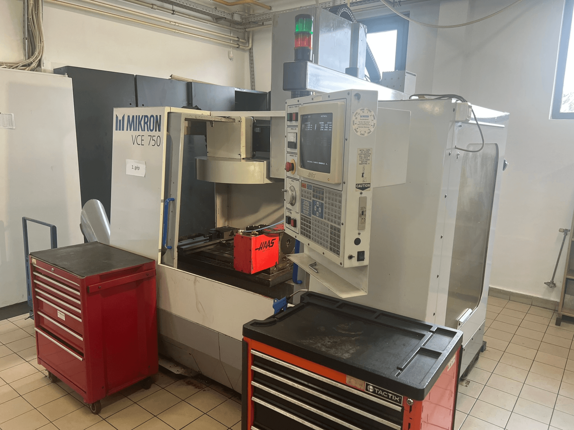 Alt-Text: "MIKRON VCE 750 Vertikal-Bearbeitungszentrum, Frontansicht mit Bedienpult und roten Werkzeugkästen, in einer geräumigen Werkstatt."