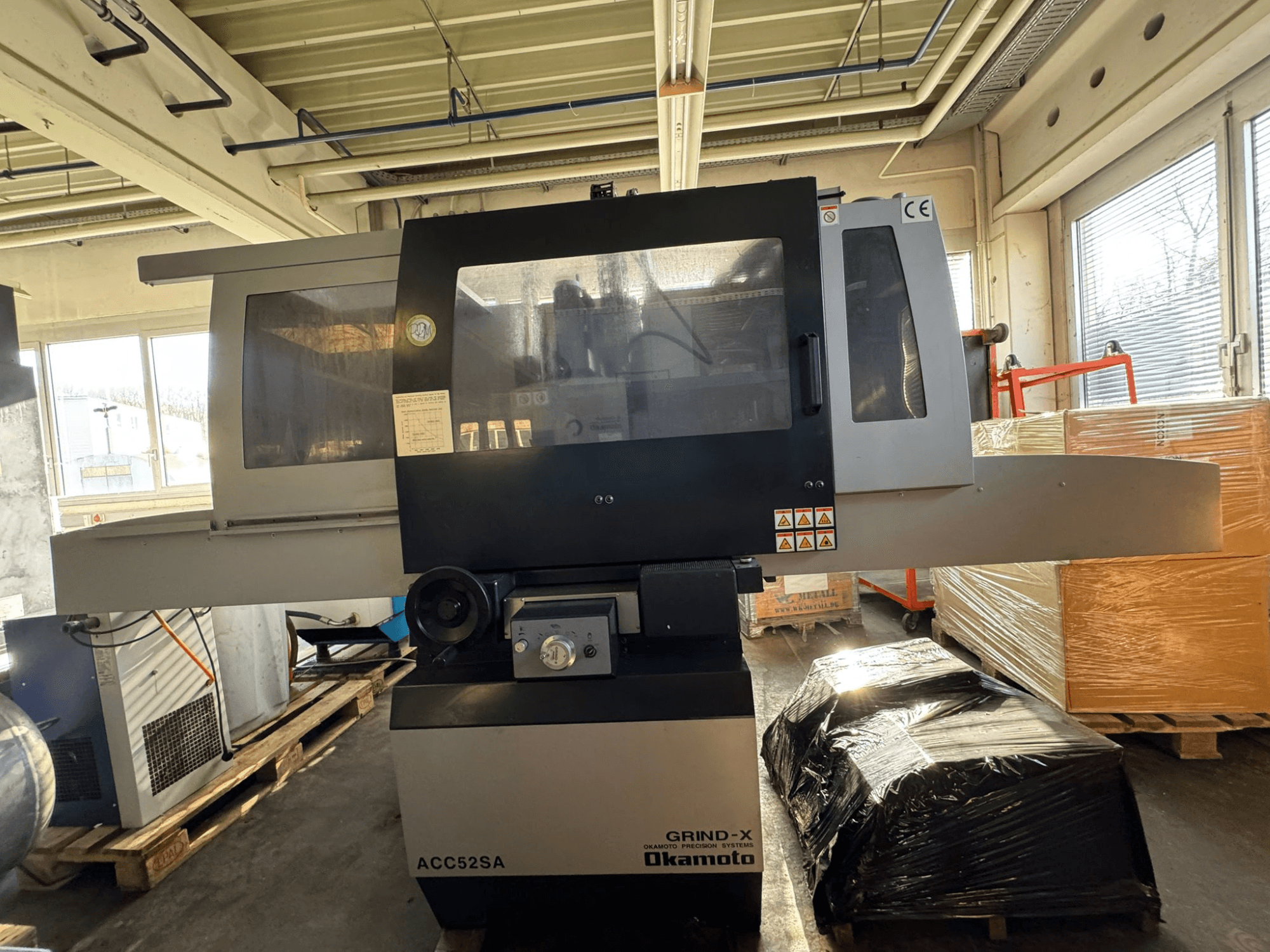 Okamoto ACC 52 SA Flachschleifmaschine in Seitenansicht, mit Bedienelementen und Beschriftungen, umgeben von Paletten und Kartons.