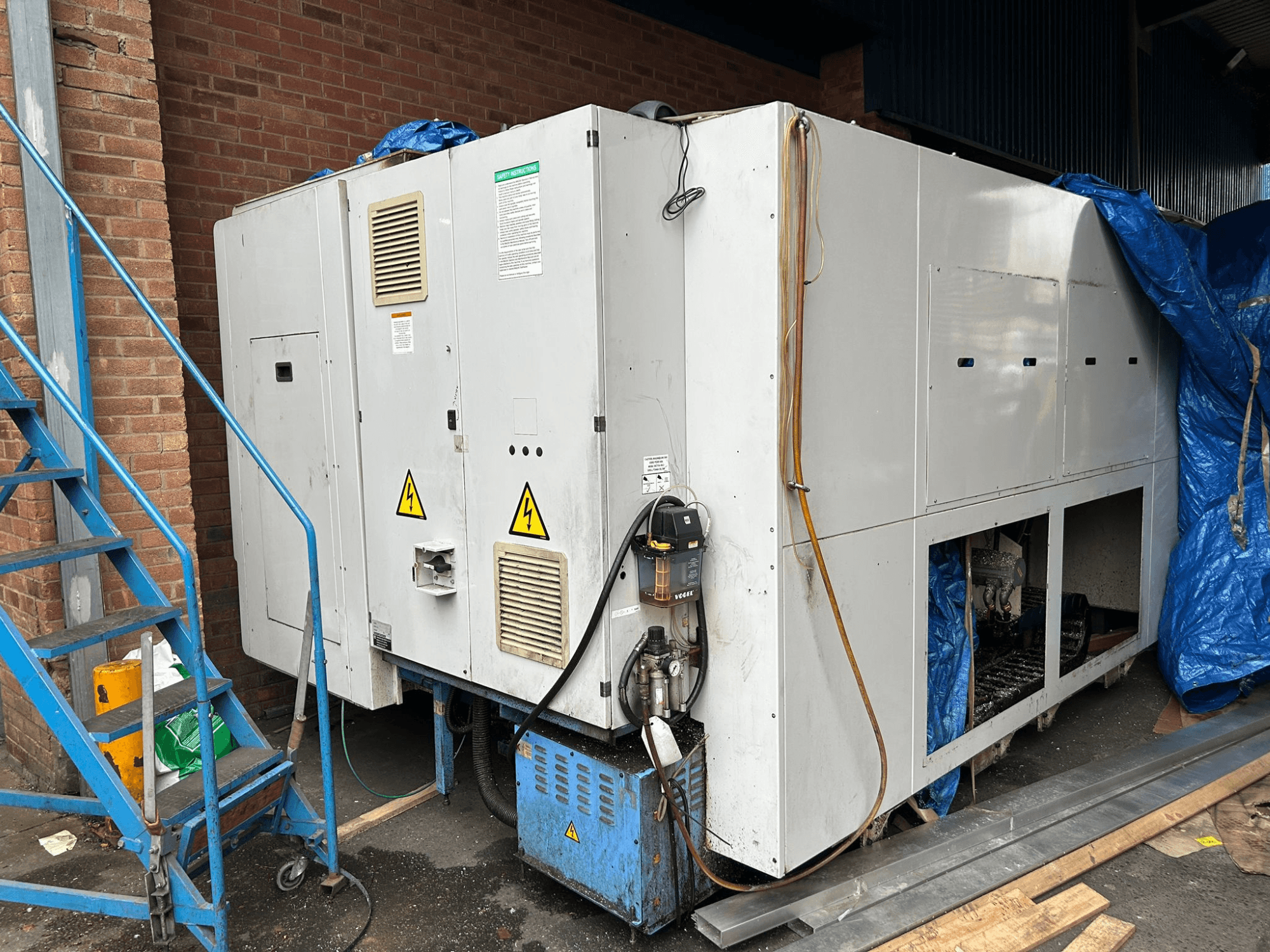 Seitenansicht eines großen Industriekühlers, Marke und Modell unklar, mit elektrischen Anschlüssen, Warnschildern und blauer Abdeckplane.