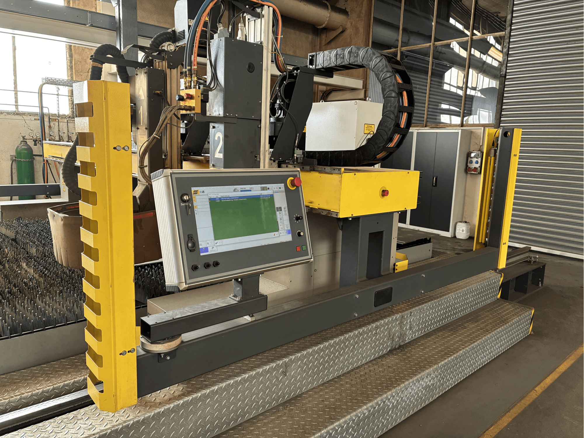 Industrielle Schneidemaschine mit Touchscreen-Bedienfeld, gelbe Schutzvorrichtung, Vorderansicht, sichtbarer Metallgitterboden.