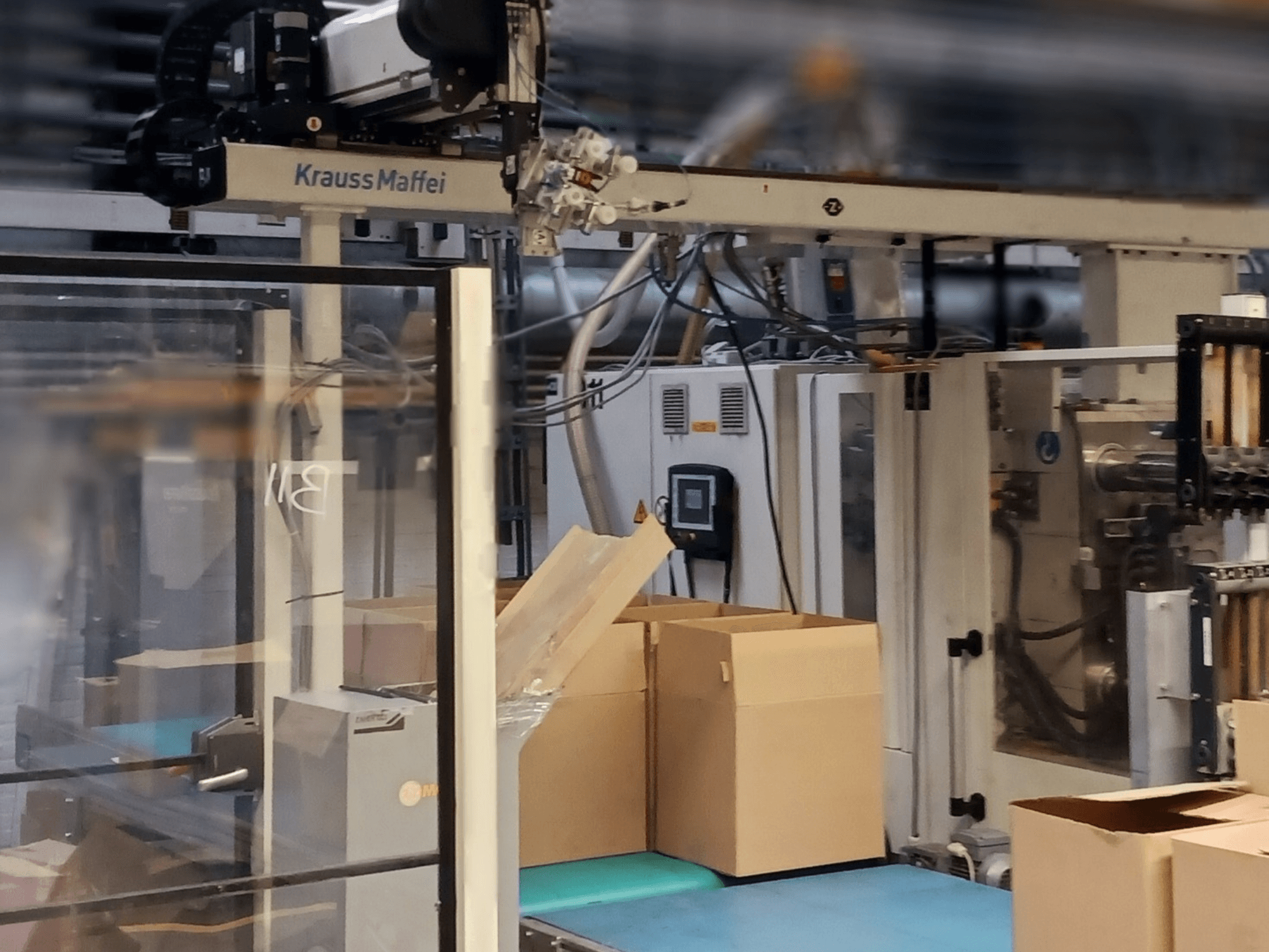 Krauss Maffei Automatisierungssystem in einer Fabrik, Frontansicht mit Kartons auf einem Förderband und Maschinenkomponenten in Betrieb.