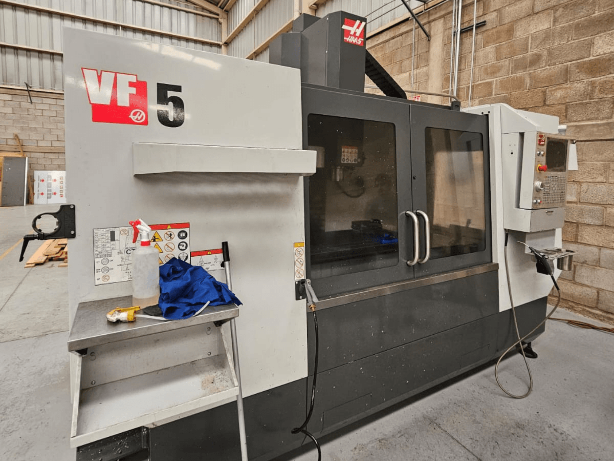 Haas VF-5 CNC-Fräsmaschine in der Vorderansicht, mit Bedienfeld, Kühlmittelflasche, blauem Tuch und Sicherheitshinweisen.