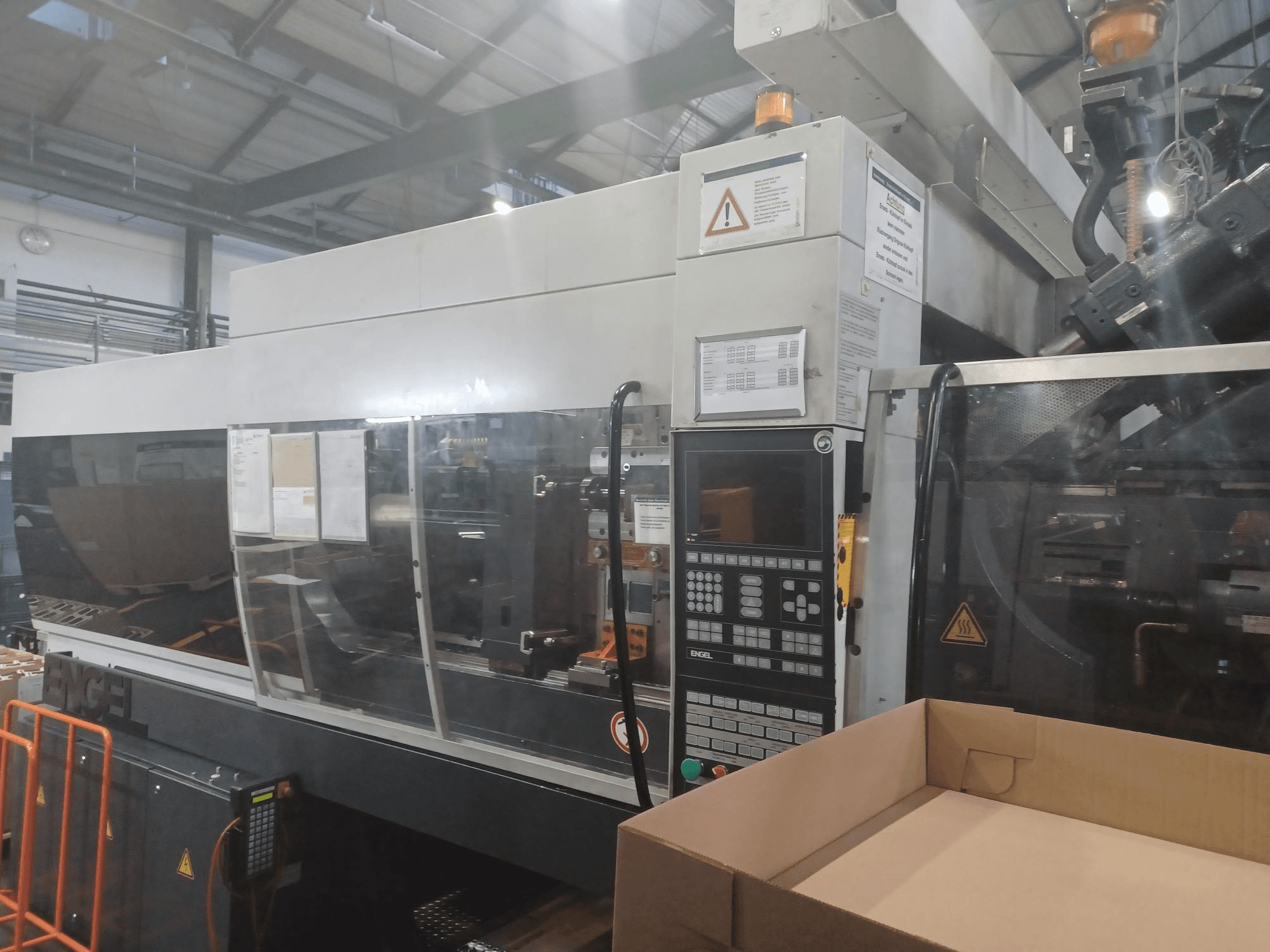 Engel-Spritzgießmaschine, Vorderansicht, mit Bedienfeld, Sicherheitsetiketten und einer leeren Schachtel in der Nähe.