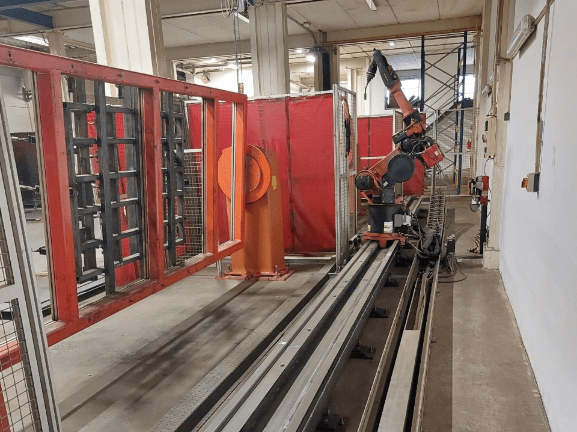 Industrieller Roboterarm in orange und schwarzer Farbe auf Metallschienen, umgeben von roten Schutzschranken, Vorderansicht.