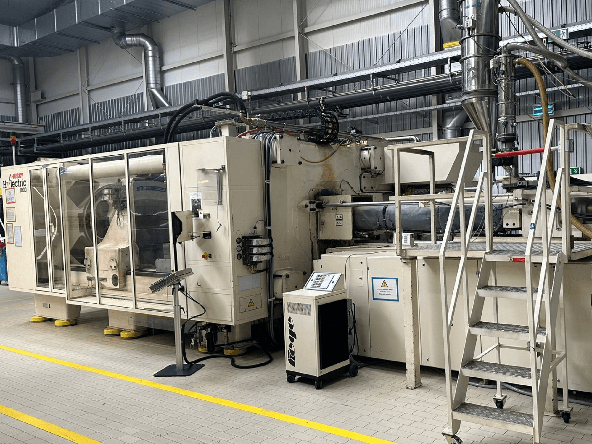 Vorderansicht einer elektrischen Spritzgießmaschine vom Typ Husky Hylelectric H1000 RS95/80 in einer Fabrikumgebung, mit Bedienfeld und Stufen.