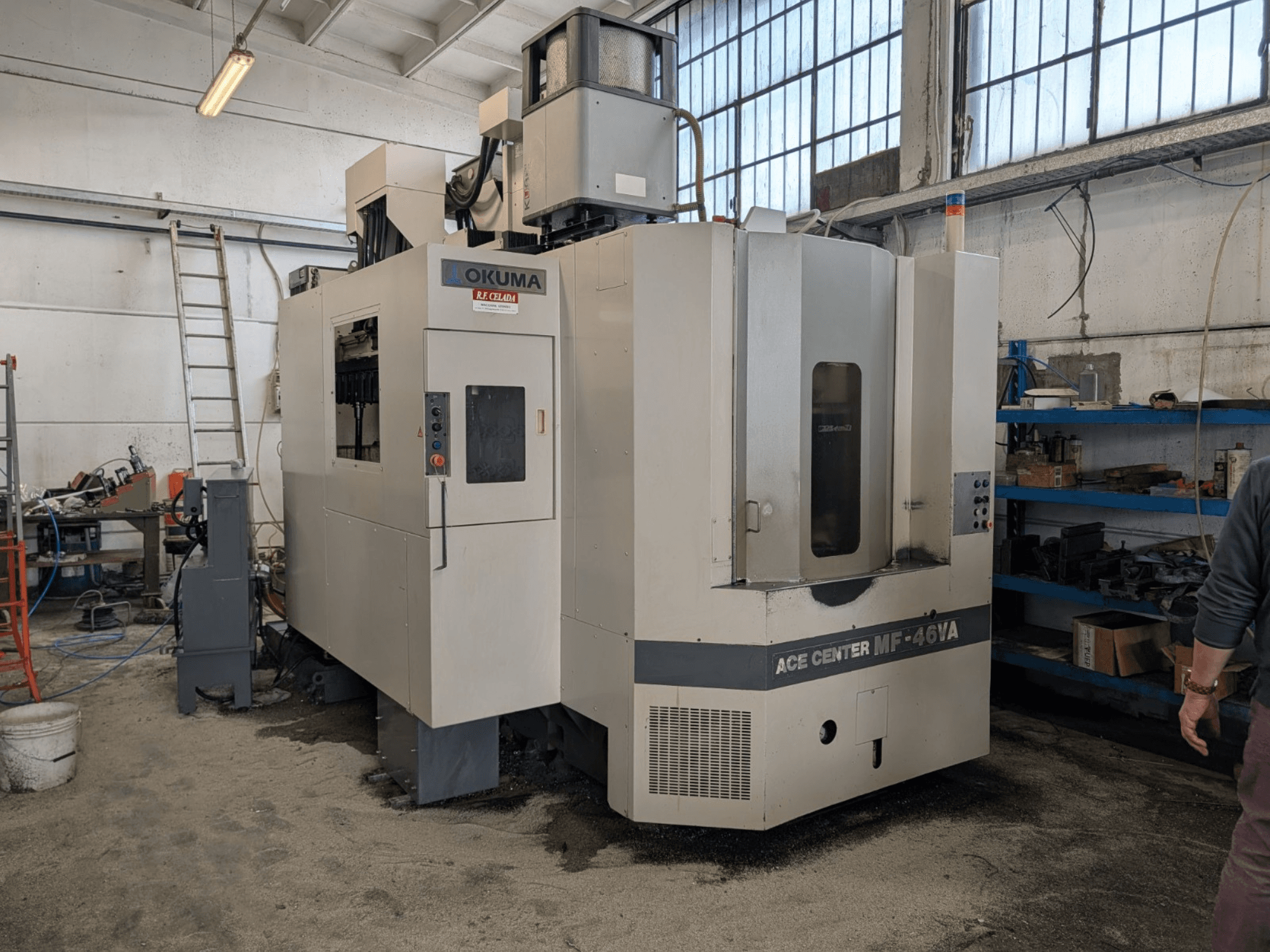 Vertikales Bearbeitungszentrum Okuma ACE CENTER MF-46VA in einer Werkstatt, Frontansicht mit Bedienfeld und umliegenden Geräten.