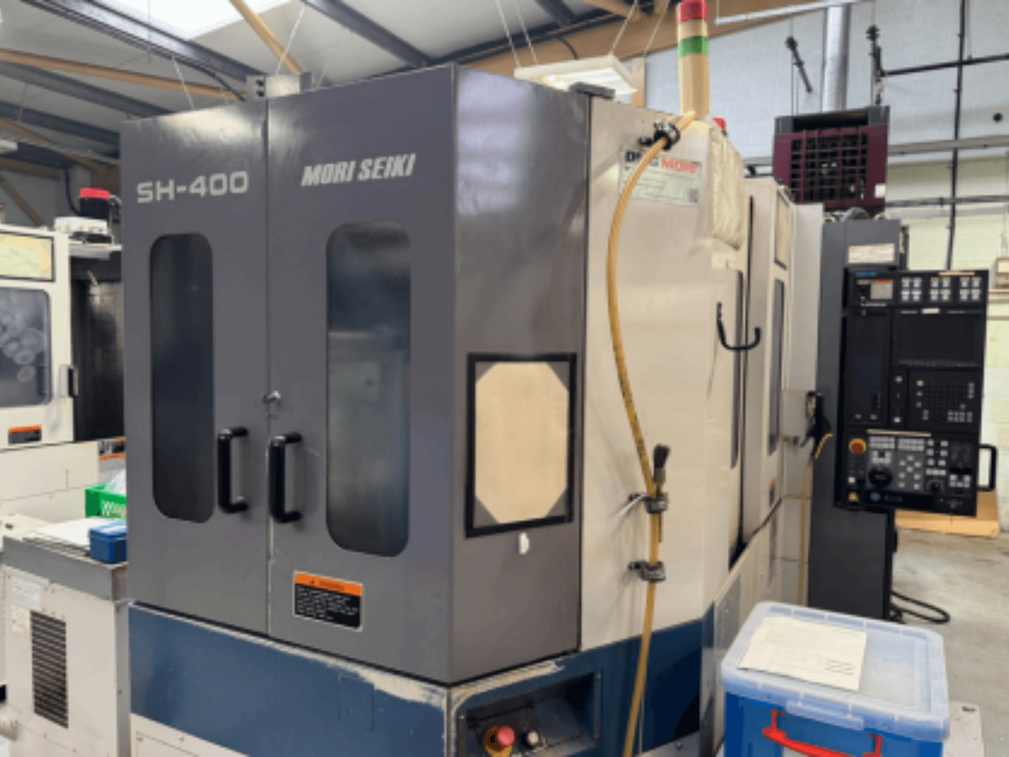 Mori Seiki SH-400 CNC-Maschine, Vorderansicht, mit Doppeltüren, Bedienfeld und Befestigungsschlauch. Blauer Lagerbehälter in der Nähe.