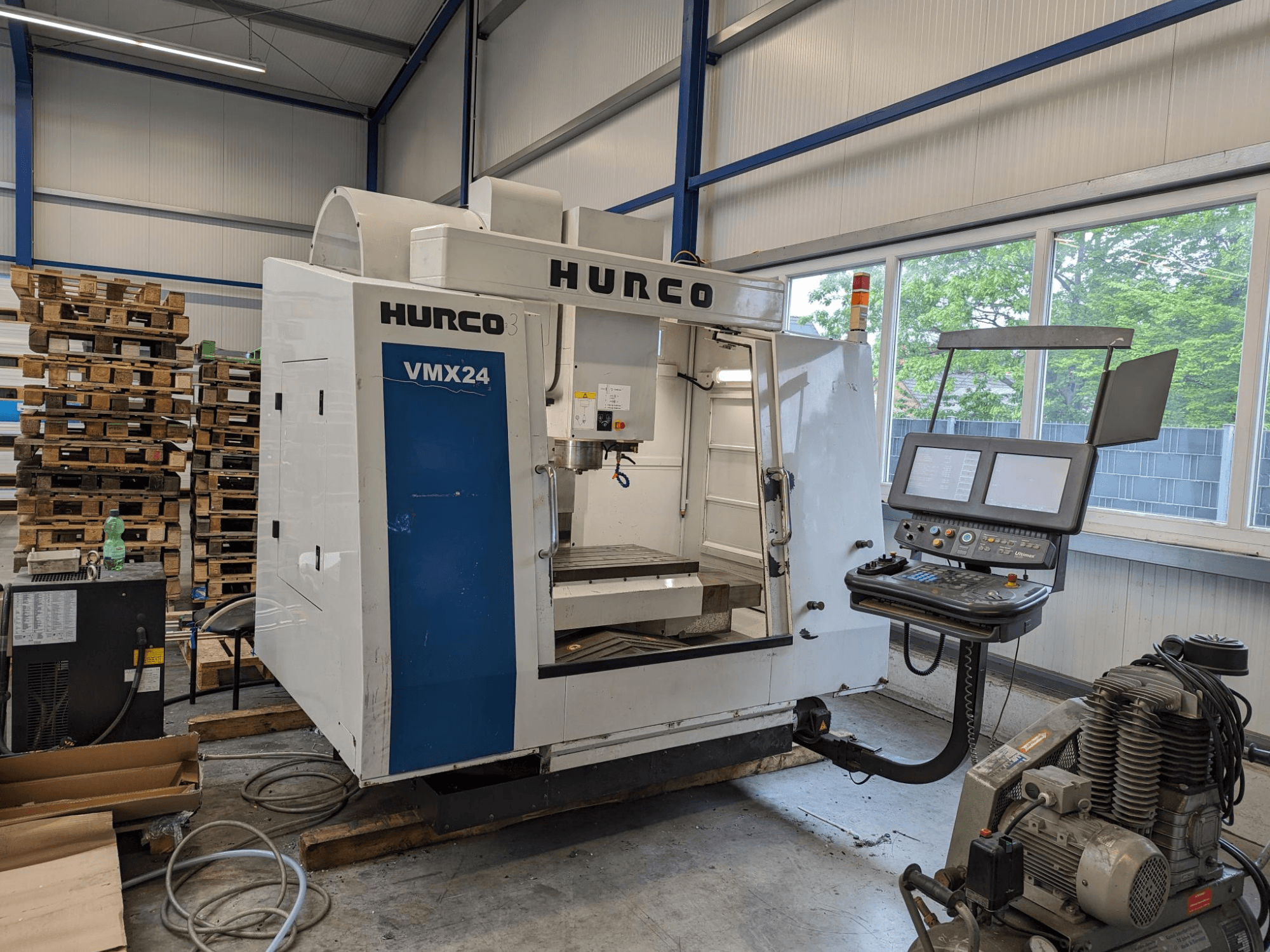 Hurco VMX24 Fräsmaschine, Vorderansicht, mit Bedienfeld und Werkzeugbereich; gestapelte Paletten im Hintergrund; industrielle Arbeitsumgebung.
