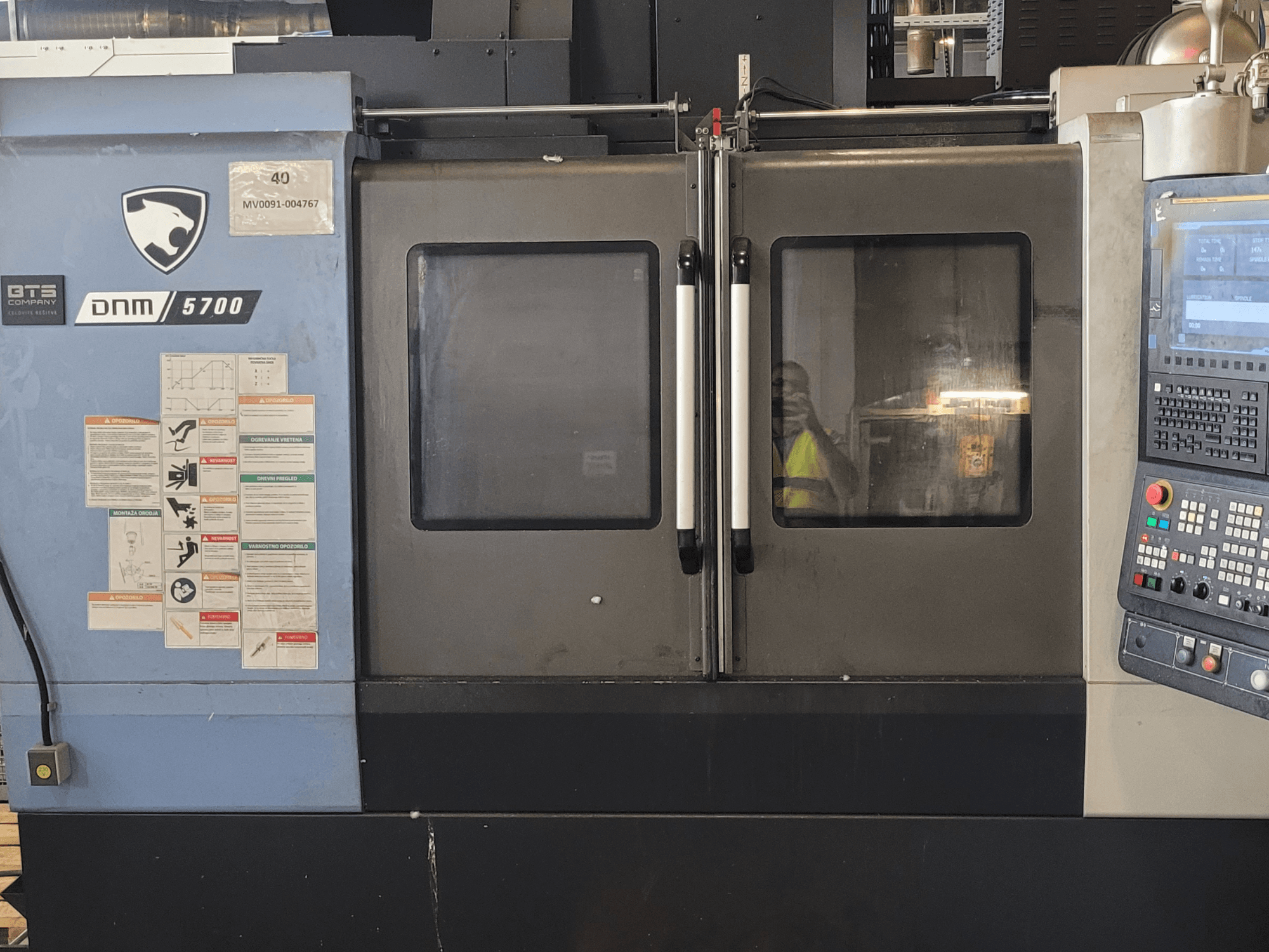 DOOSAN DNM 5700 Vertikal-Bearbeitungszentrum, Vorderansicht mit Bedienfeld, Sicherheitsaufklebern und leicht angelehnten Türen.