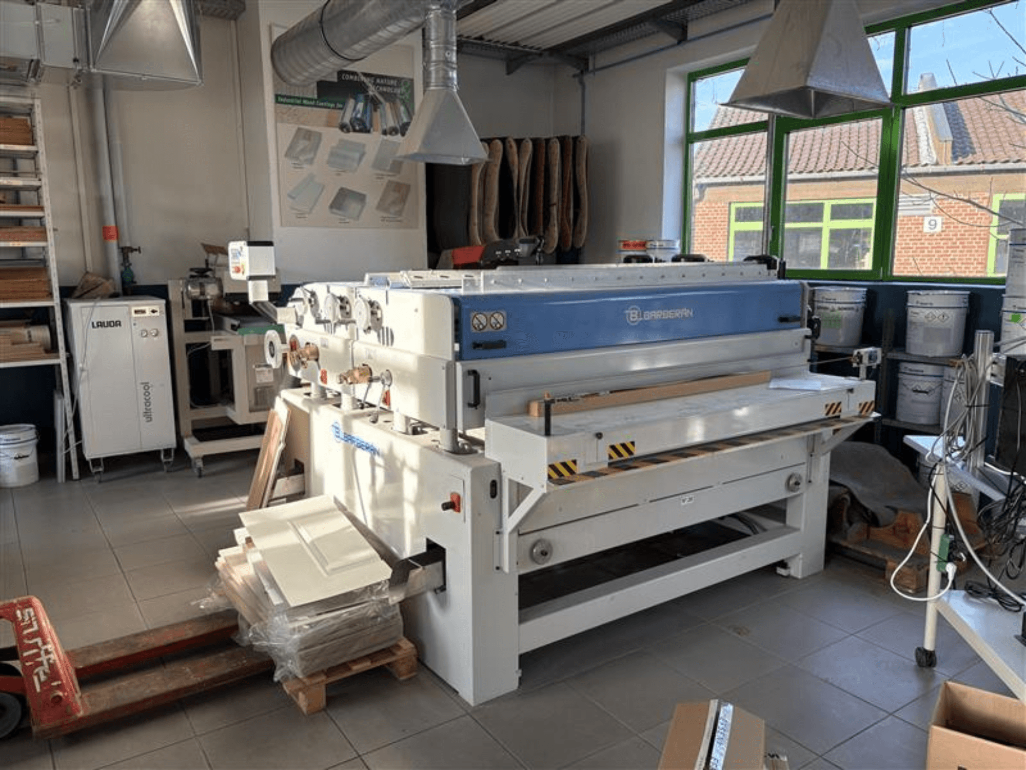 Blaserbn Holzbearbeitungsmaschine in einer Werkstatt, Vorderansicht; mit Holzplatten auf einer Palette und modernen Geräten im Hintergrund.