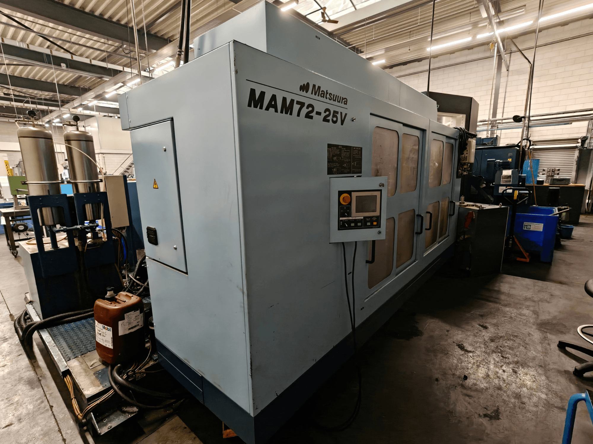 Matsuura MAM72-25V CNC-Maschine, Seitenansicht, mit Bedienfeld, zwei Kühlmitteltanks und Arbeitsbereich in einer industriellen Umgebung.