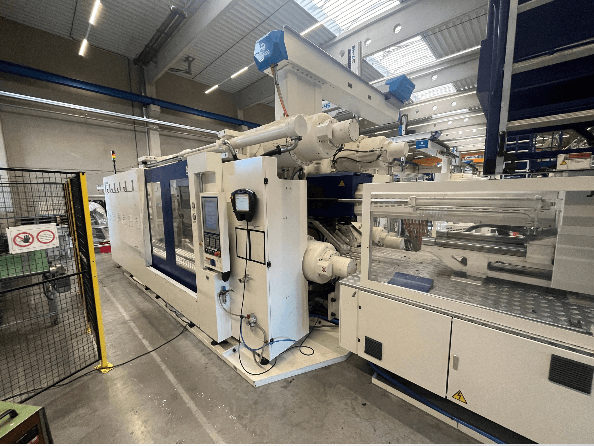 Spritzgießmaschine in Weiß und Blau, Vorderansicht, mit Bedienfeld, Roboterarm und Sicherheitsschildern.