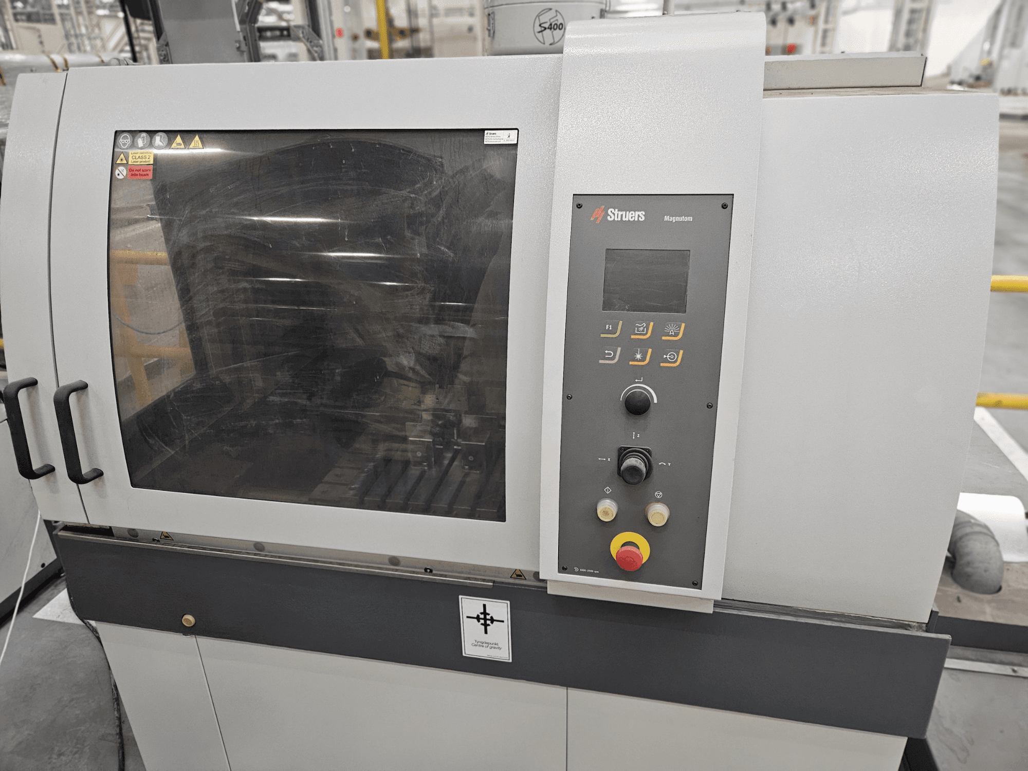 Vorderansicht der Struers Magpentor Maschine mit Bedienfeld, Tasten und einem großen Sichtfenster. Technische Etiketten sichtbar.