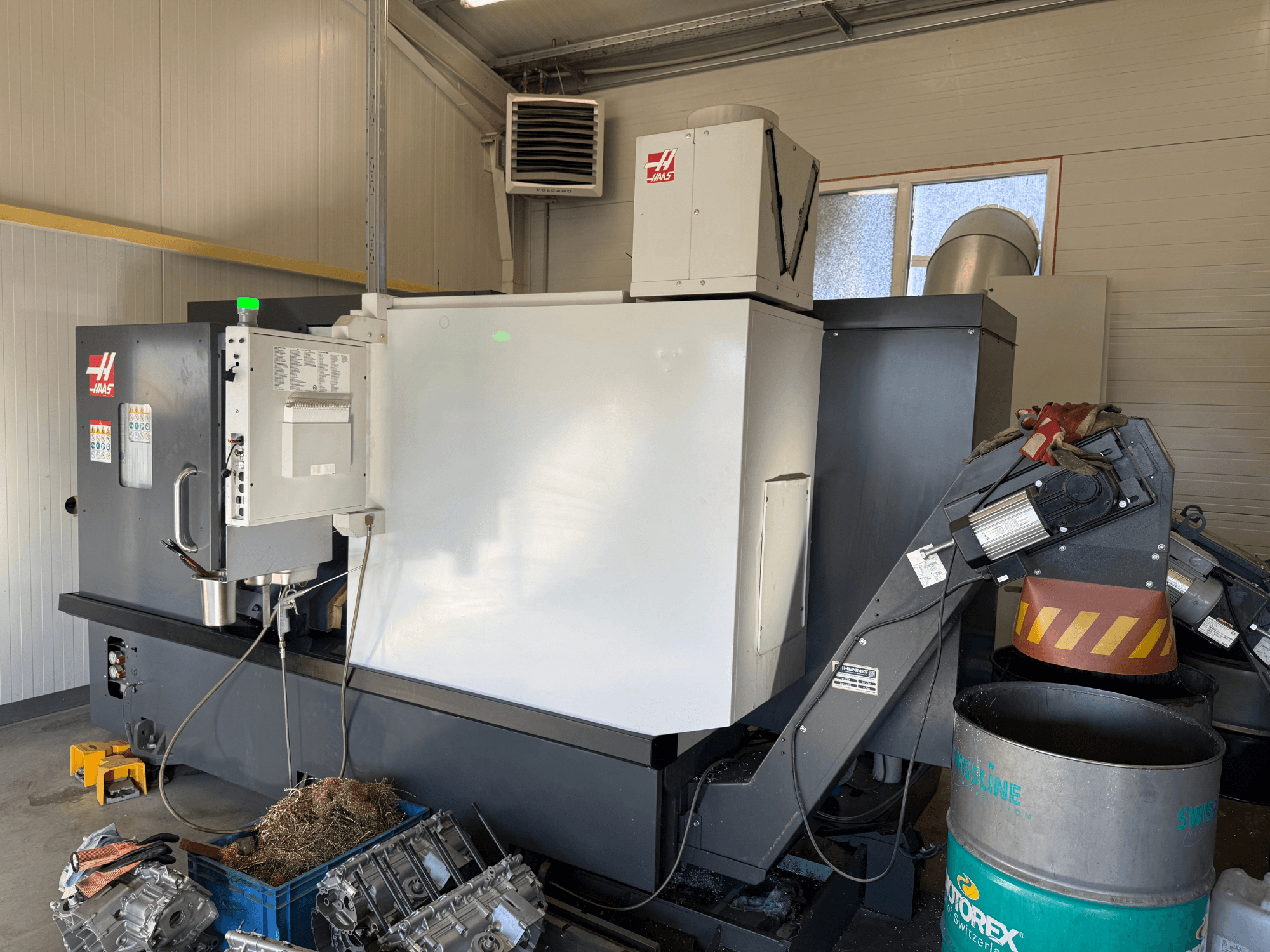 Haas CNC-Maschine mit Bedienfeld, Seitenansicht; sichtbare Kühlmittel- und Ölleitungen; Metallabfälle in blauer Tonne; Industrieumgebung.