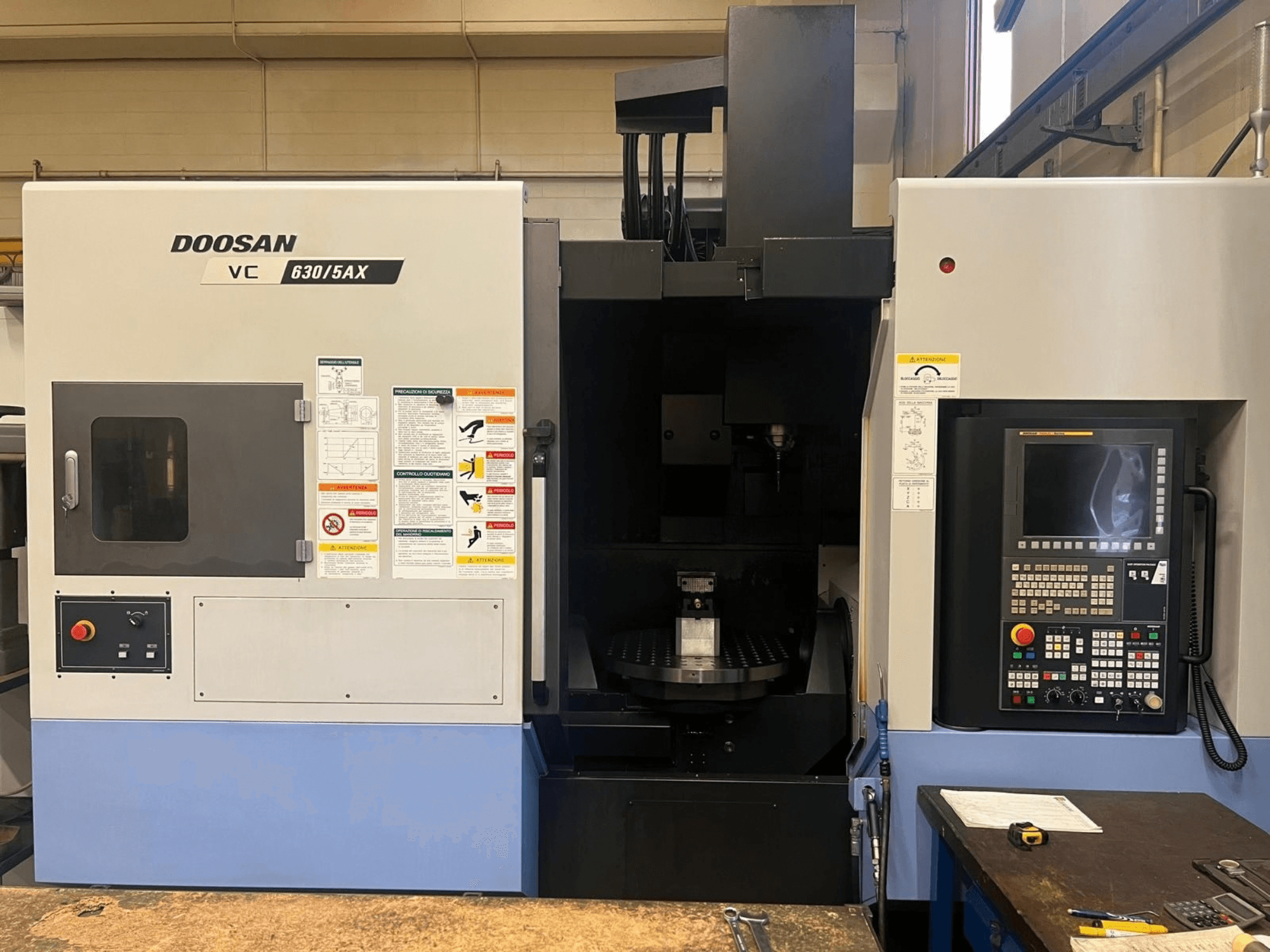 Vorderansicht eines vertikalen Bearbeitungszentrums vom Typ DOOSAN VC 630/5AX, auf der das Bedienfeld und der Spindelbereich mit gut sichtbaren Sicherheitsaufklebern zu sehen sind.