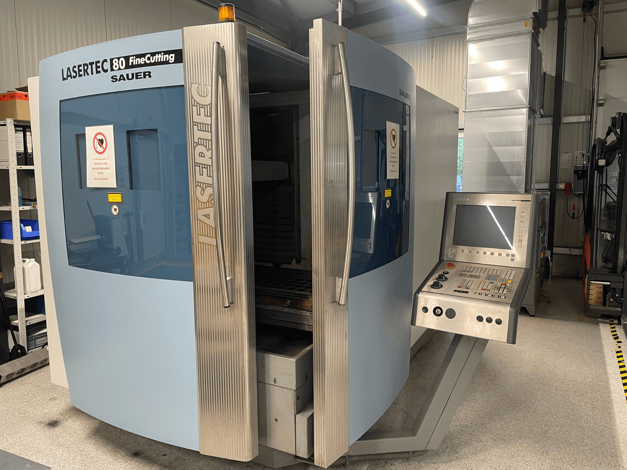 Vorderansicht der Blechbearbeitungsmaschine „DMG Sauer Lasertec80 FineCutting“ mit Bedienfeld, geöffneten Türen und sichtbaren Sicherheitshinweisen.