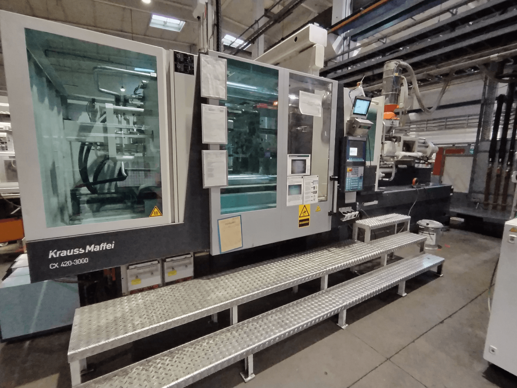 Krauss Maffei CX 420-3000 Spritzgießmaschine, Seitenansicht, mit Bedienpult und Sicherheitsschildern sichtbar, auf Metallstufen aufgestellt.