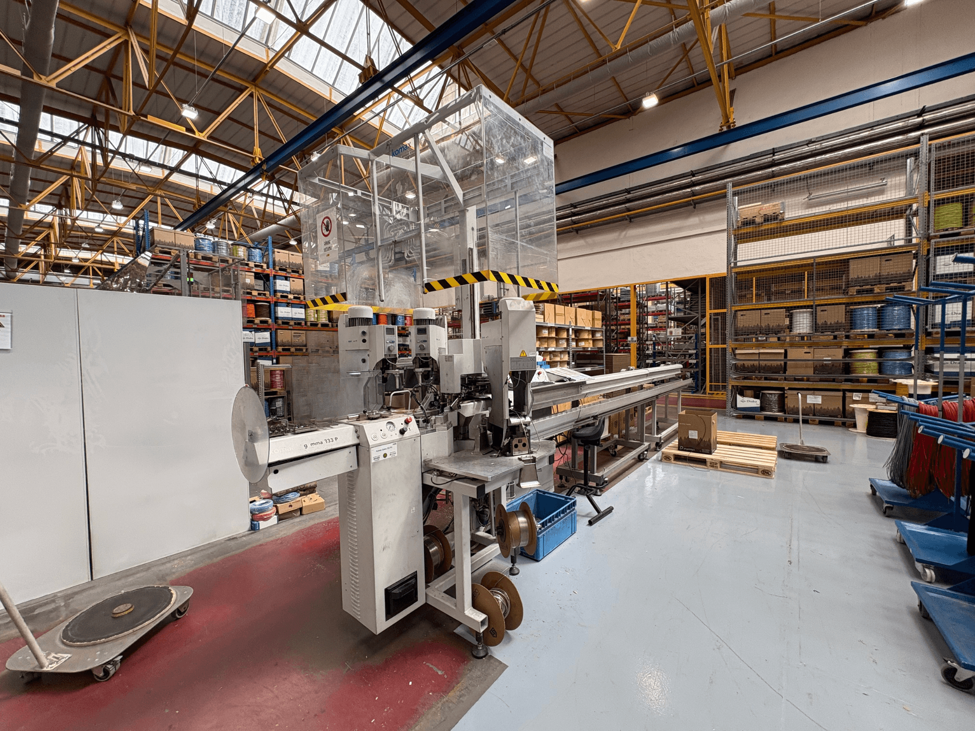 Werkzeugmaschine KOMAX GAMMA 333 PC in einem Lager, Seitenansicht mit Bedienelementen, Förderanlage und umliegenden Regalen.