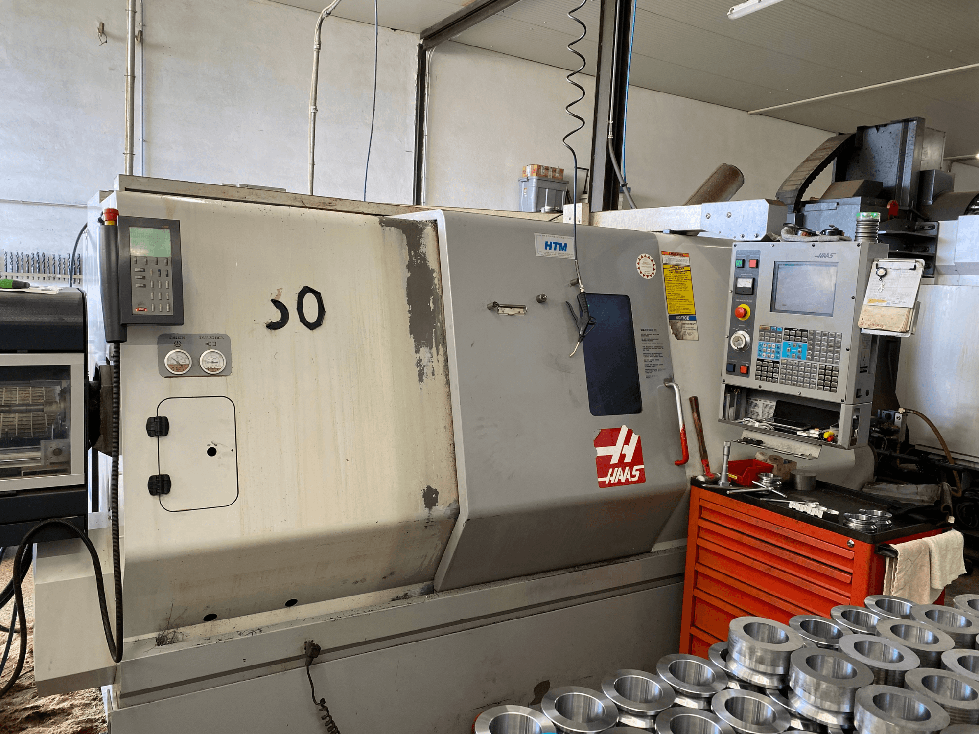 Haas CNC-Maschine mit Bedienfeld, Vorderansicht, mit Bedientasten, Messgeräten und daneben gestapelten Metallteilen.