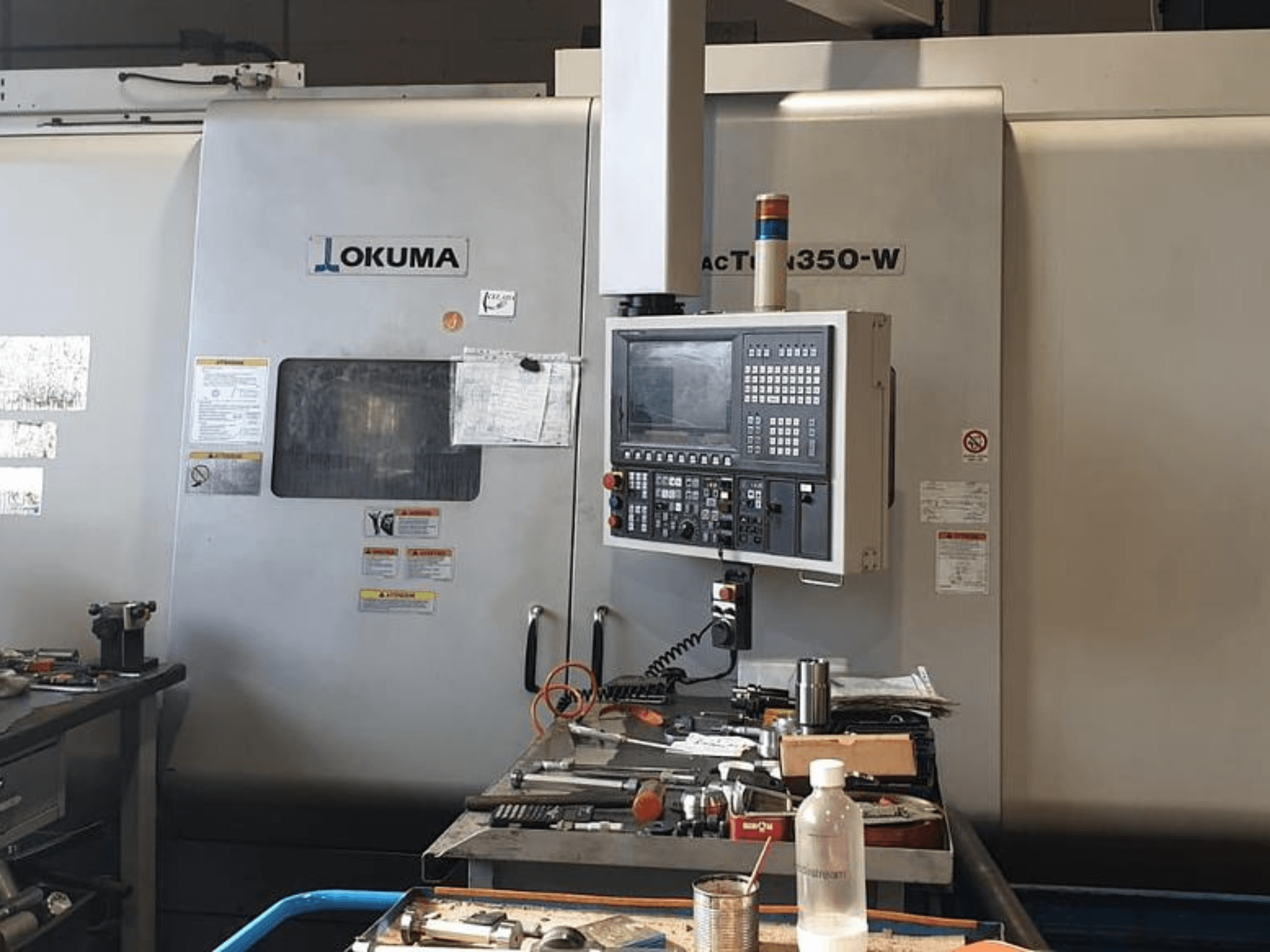 Okuma MAC TURN 350-W Dreh-Fräszentrum, Vorderansicht mit Bedienpult und Arbeitsfläche mit Werkzeugen und einer Flasche vollgestopft.