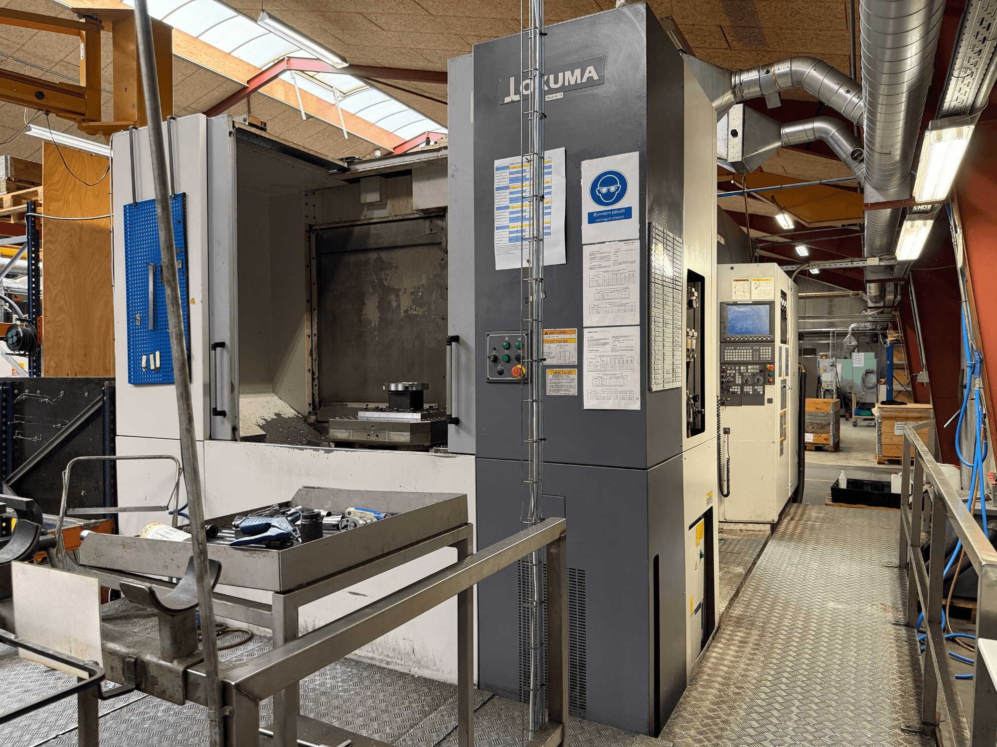 Horizontales Bearbeitungszentrum Okuma MA-600HB, Vorderansicht, mit Werkzeugen auf einem Metalltisch und der Maschinensteuerung.