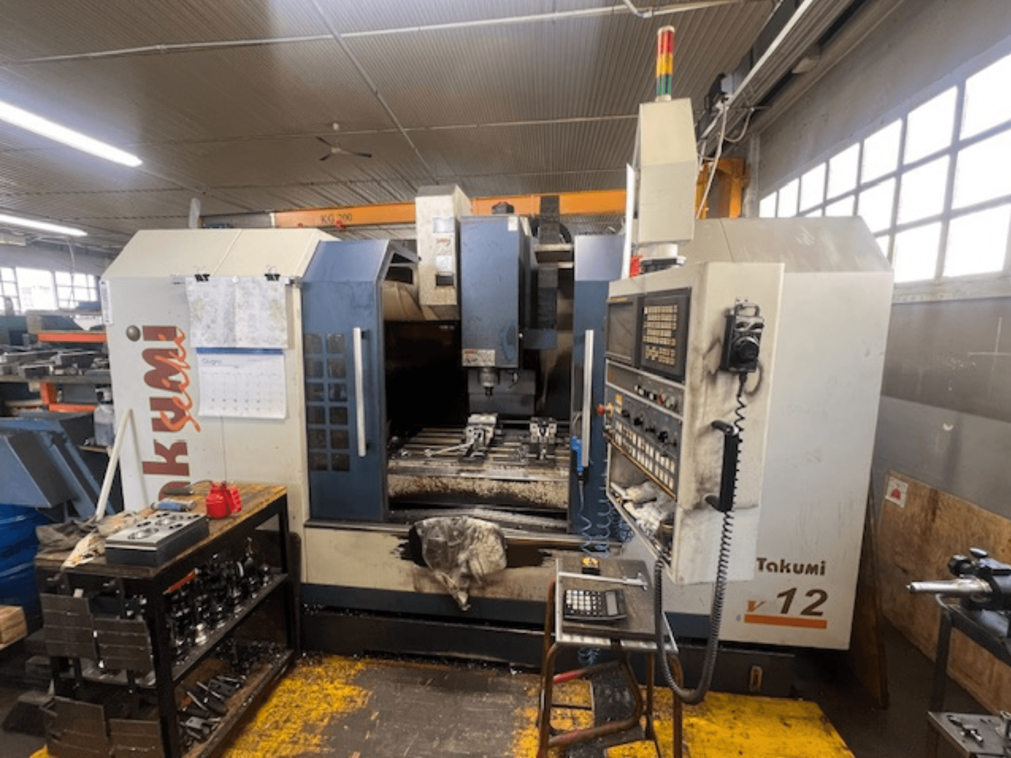 Takumi V12 CNC-Fräsmaschine in der Seitenansicht, mit Bedienfeld, Schneidbereich und Werkzeugen auf der Arbeitsstation.