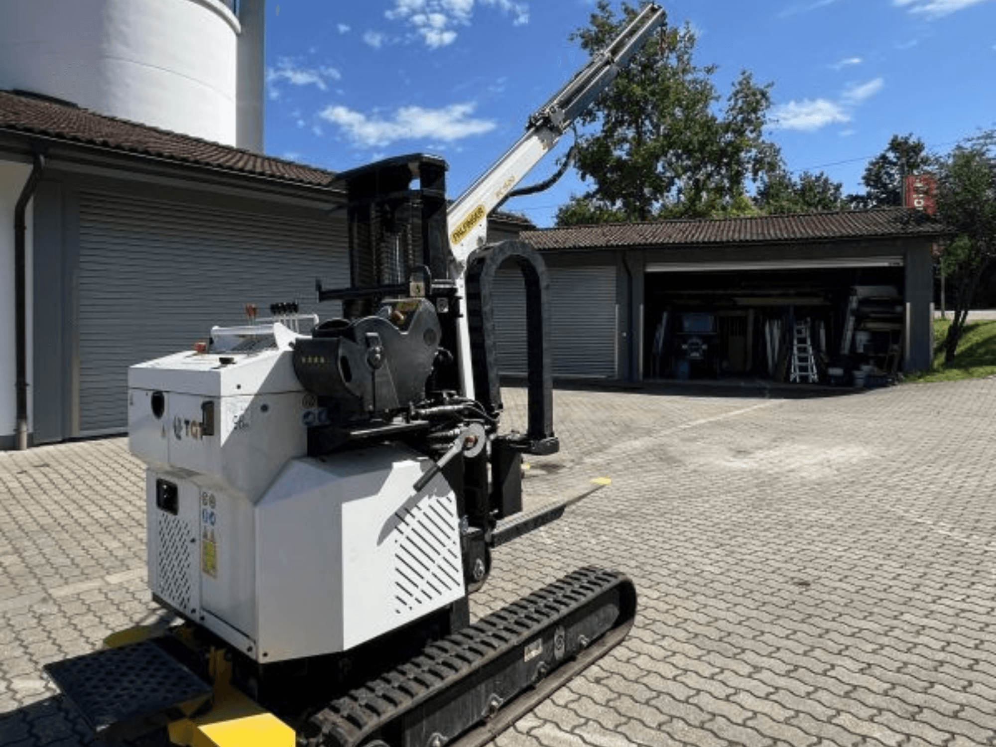 TGI-Bau-Minikran, Seitenansicht, mit Raupen und montiertem Ausleger, abgestellt auf einer gemauerten Fläche neben einer Garage.