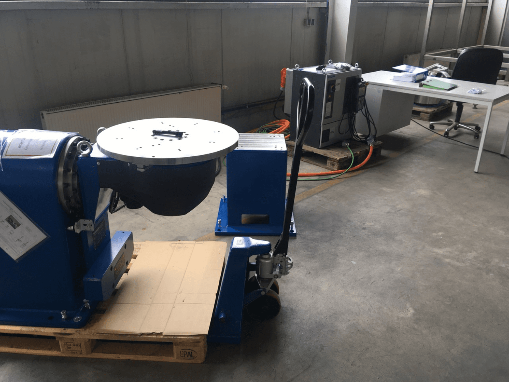 Blaues Industriegerät mit kreisförmigem Sockel und Bedienfeld, von vorne gesehen, auf einer Holzpalette in einer Büroumgebung.