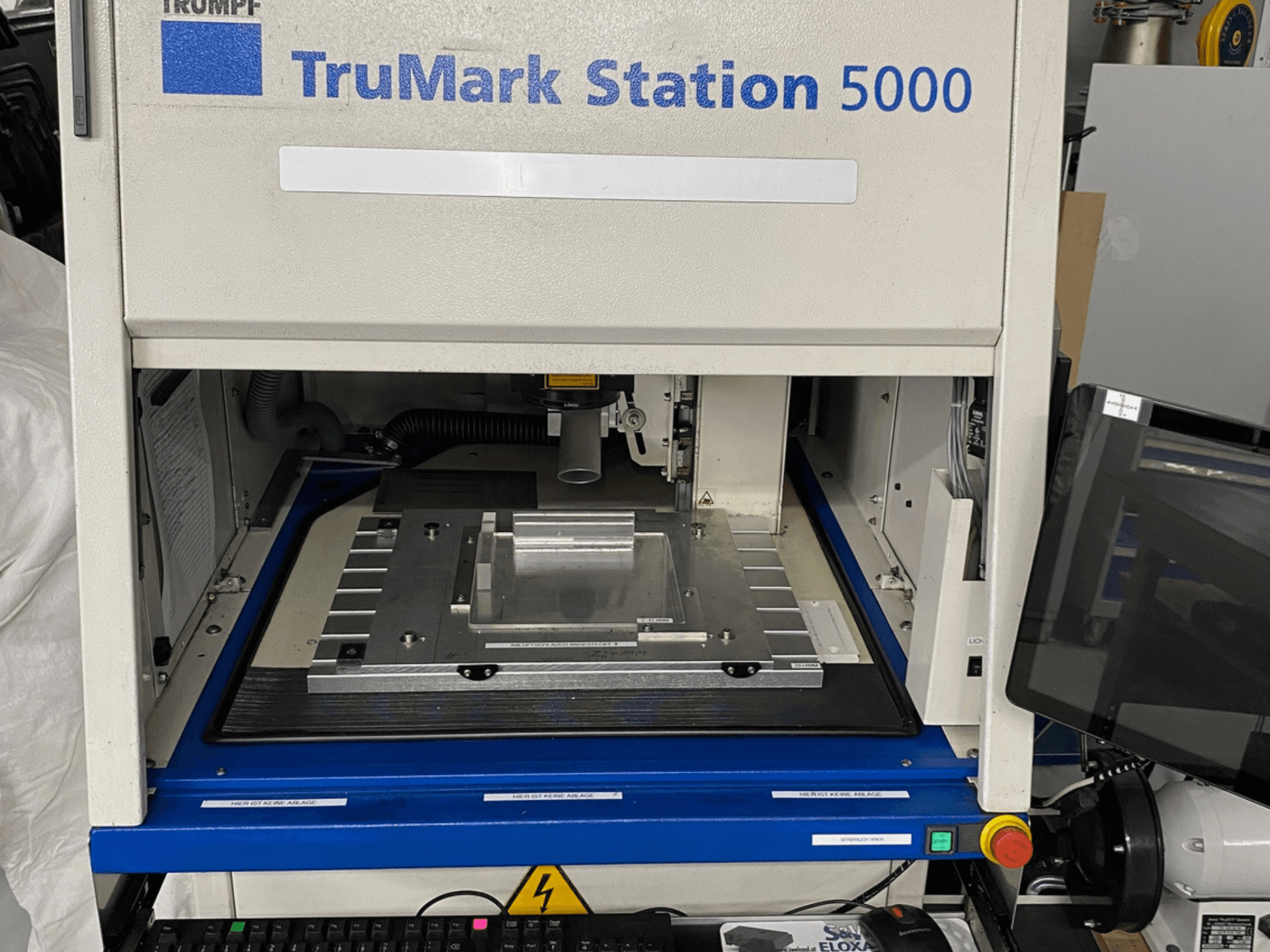 Die Vorderansicht der TruMark Station 5000 Lasermarkiermaschine zeigt den Arbeitstisch, die Bedienelemente und den Überwachungsbildschirm.