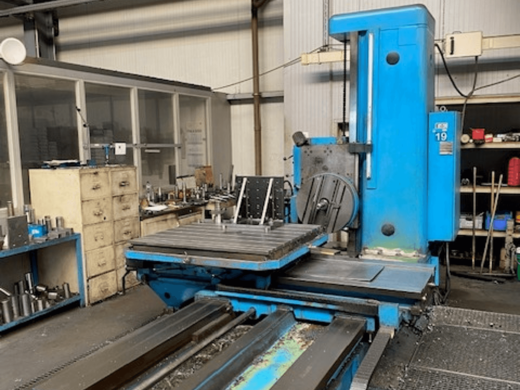 Blaue Fräsmaschine in Vorderansicht, Modellnummer 19, auf dem Boden einer Werkstatt mit Werkzeugen und einem Lagerschrank im Hintergrund.