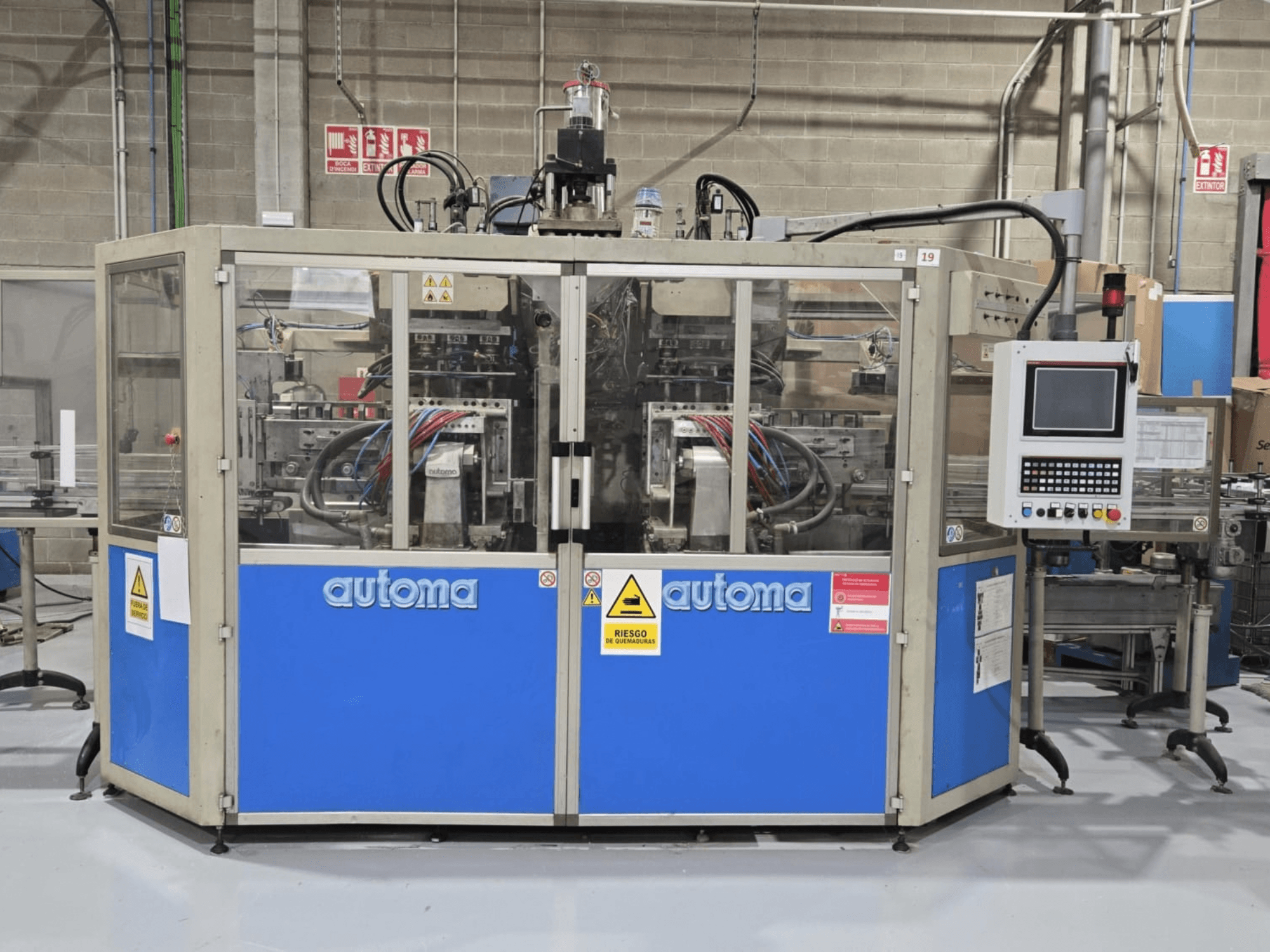 Vorderansicht der AUTOMA APEX AT500D-Blasformmaschine mit Bedienfeld und verschiedenen Schläuchen in einer Fabrikumgebung.