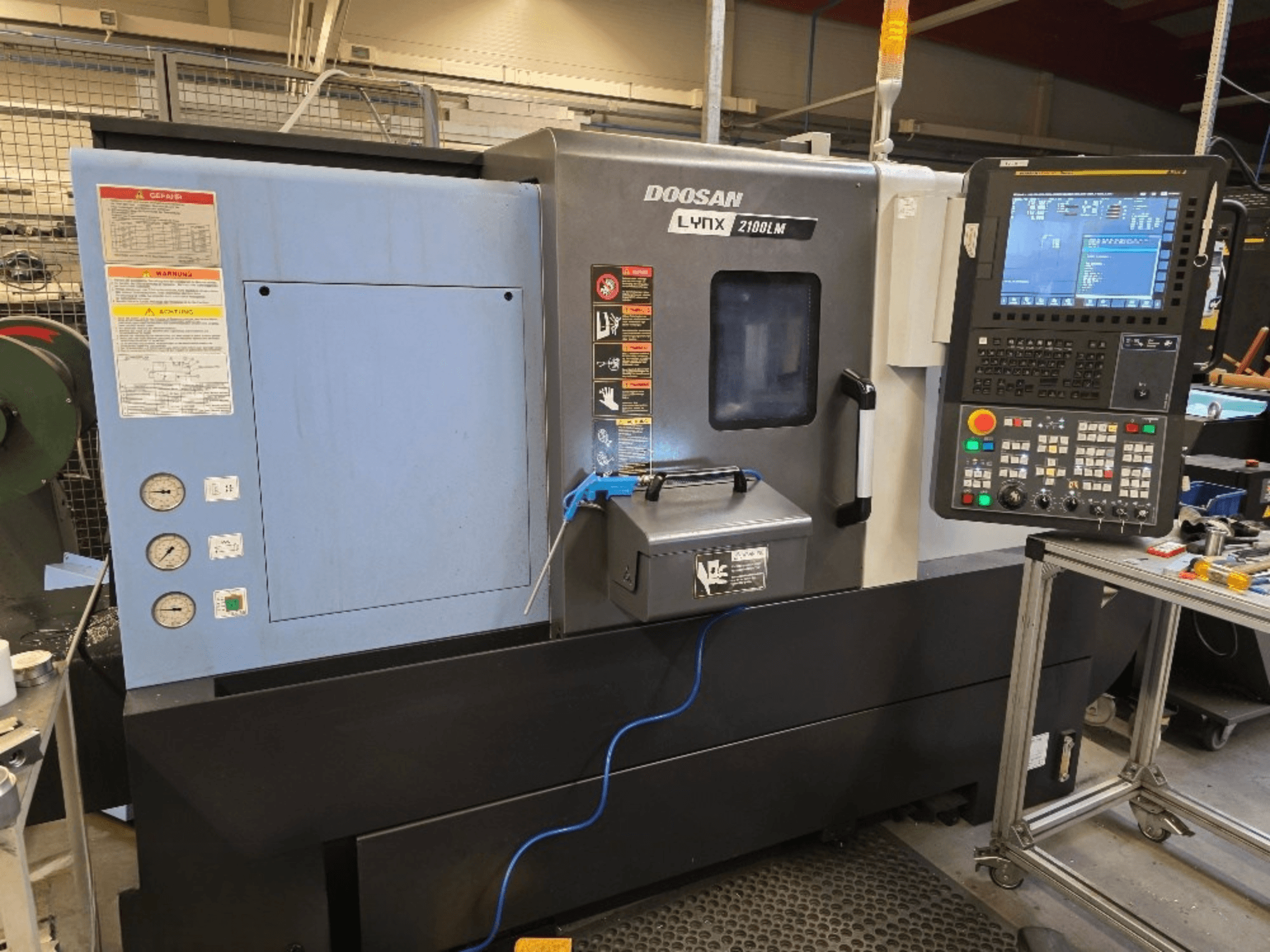 Doosan Lynx 2100LM CNC-Drehmaschine, Vorderansicht, mit Bedienfeld, Manometern und Sicherheitswarnungen.